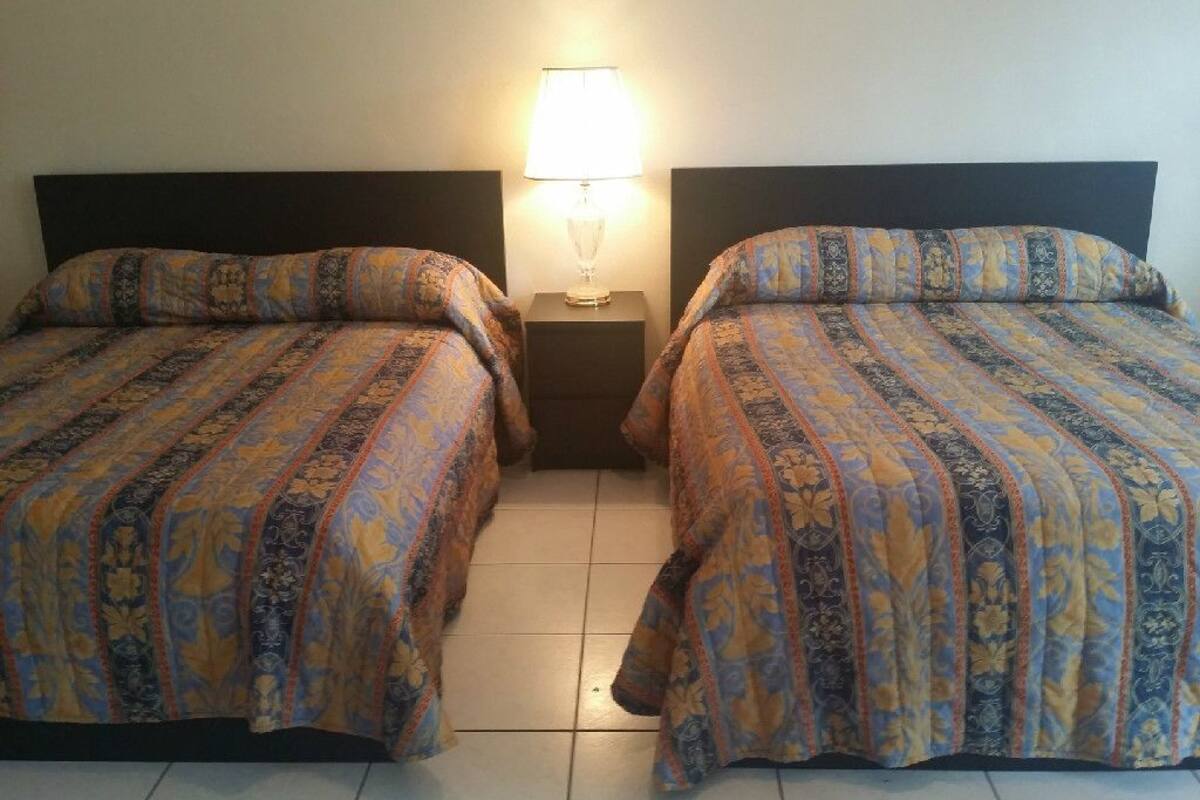 Two beds with patterned bedding, a nightstand with a lamp, and a tiled floor.