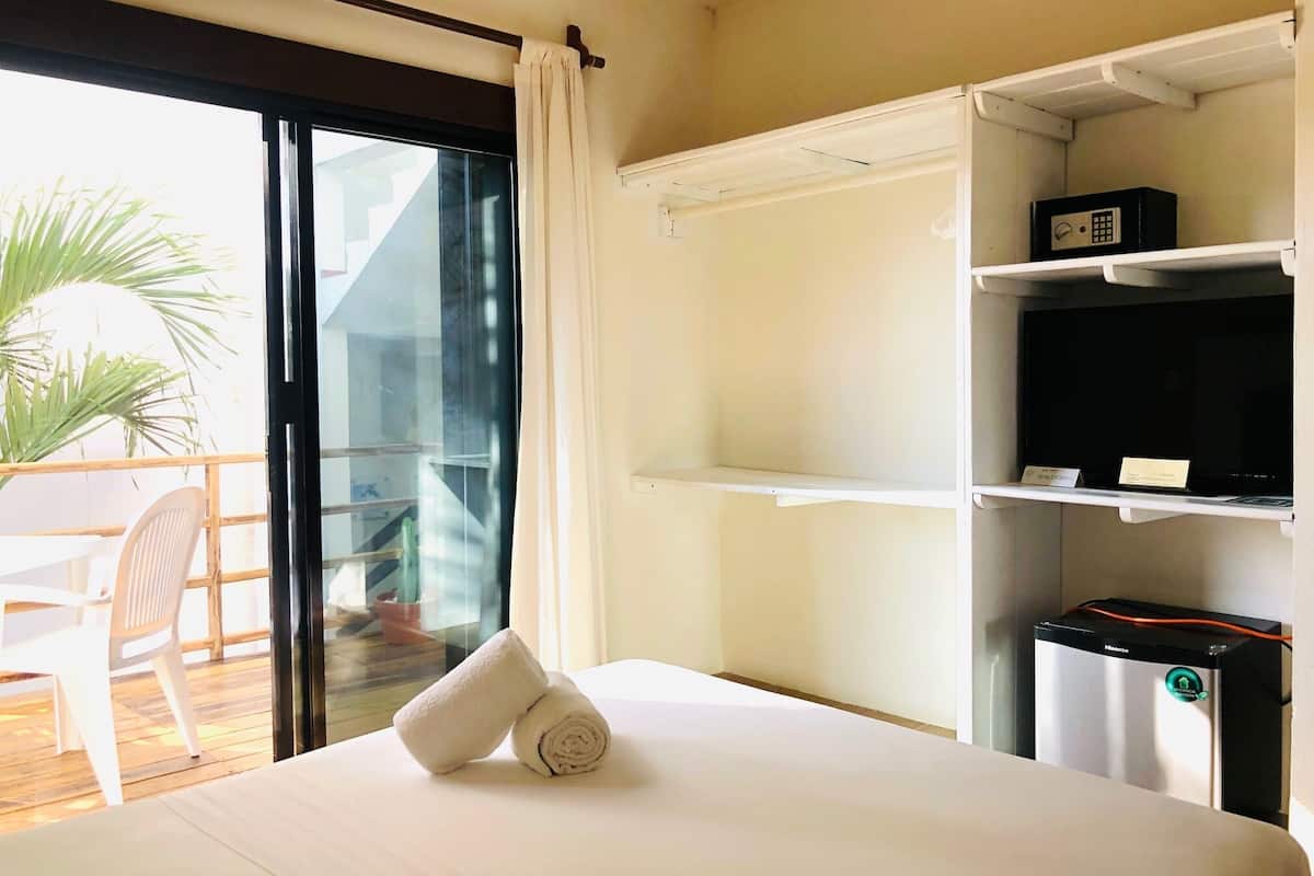 A hotel room with a bed, a TV, a minibar, and a balcony with a chair and plant.