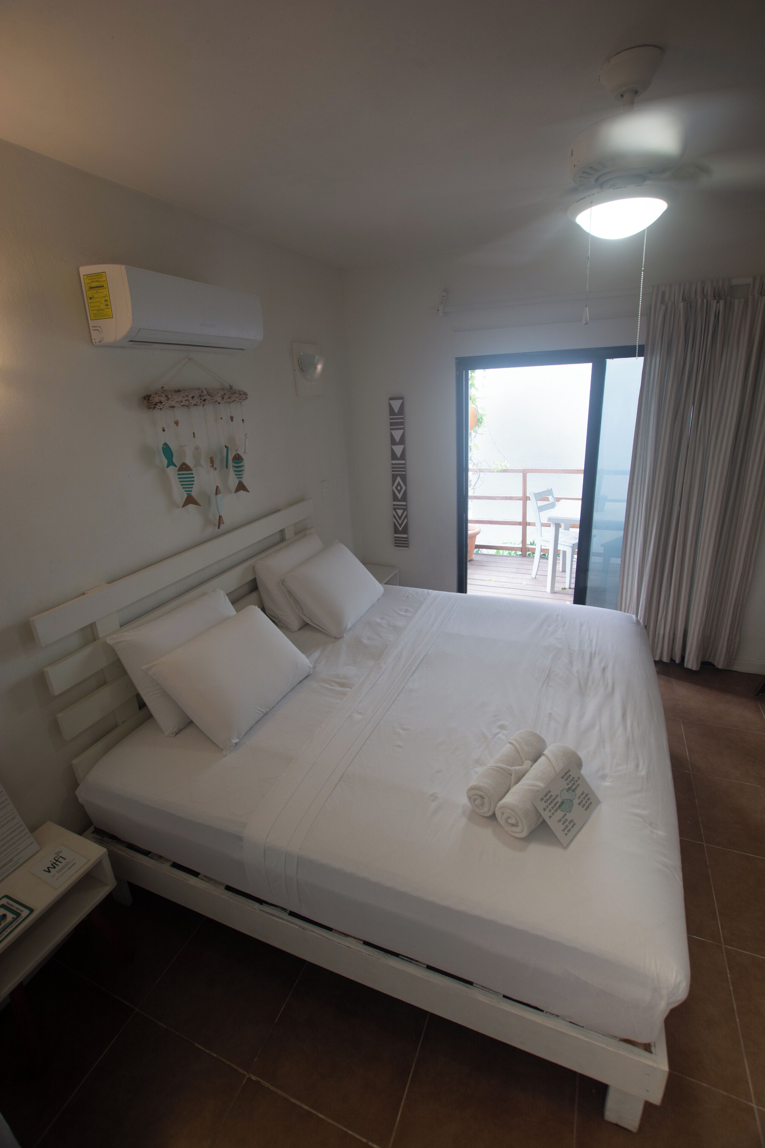 Un lit bien fait, avec du linge de lit blanc et des oreillers, un ventilateur de plafond et une vue sur l’extérieur grâce à une porte-fenêtre coulissante.