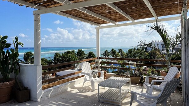 Un espacio de estar al aire libre cubierto con sillas de plástico blanco y una mesa, con vista a una playa con palmeras y el océano.