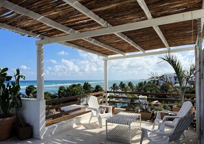 A covered outdoor seating area with white plastic chairs and a table, overlooking a beach with palm trees and the ocean. - Hotel Boutique Pachamama (Mahahual)