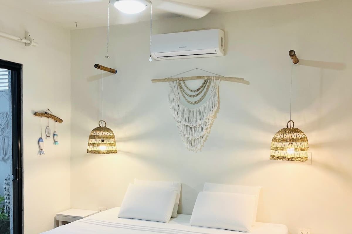 A hotel room with a large bed, a ceiling fan, an air conditioning unit, and decorative macramé wall hangings.