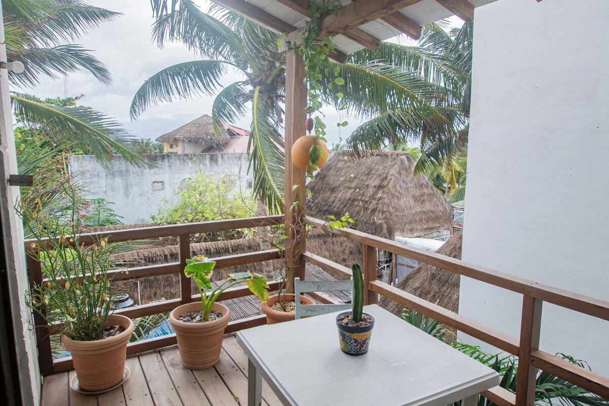 A wooden deck with a table and chair, surrounded by potted plants and a view of palm trees and a thatched-roof building.