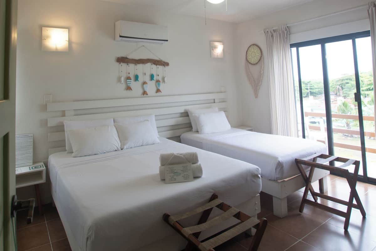 A hotel room with two beds, a ceiling fan, an air conditioning unit, a small table, and a view of the outdoors through a sliding glass door.