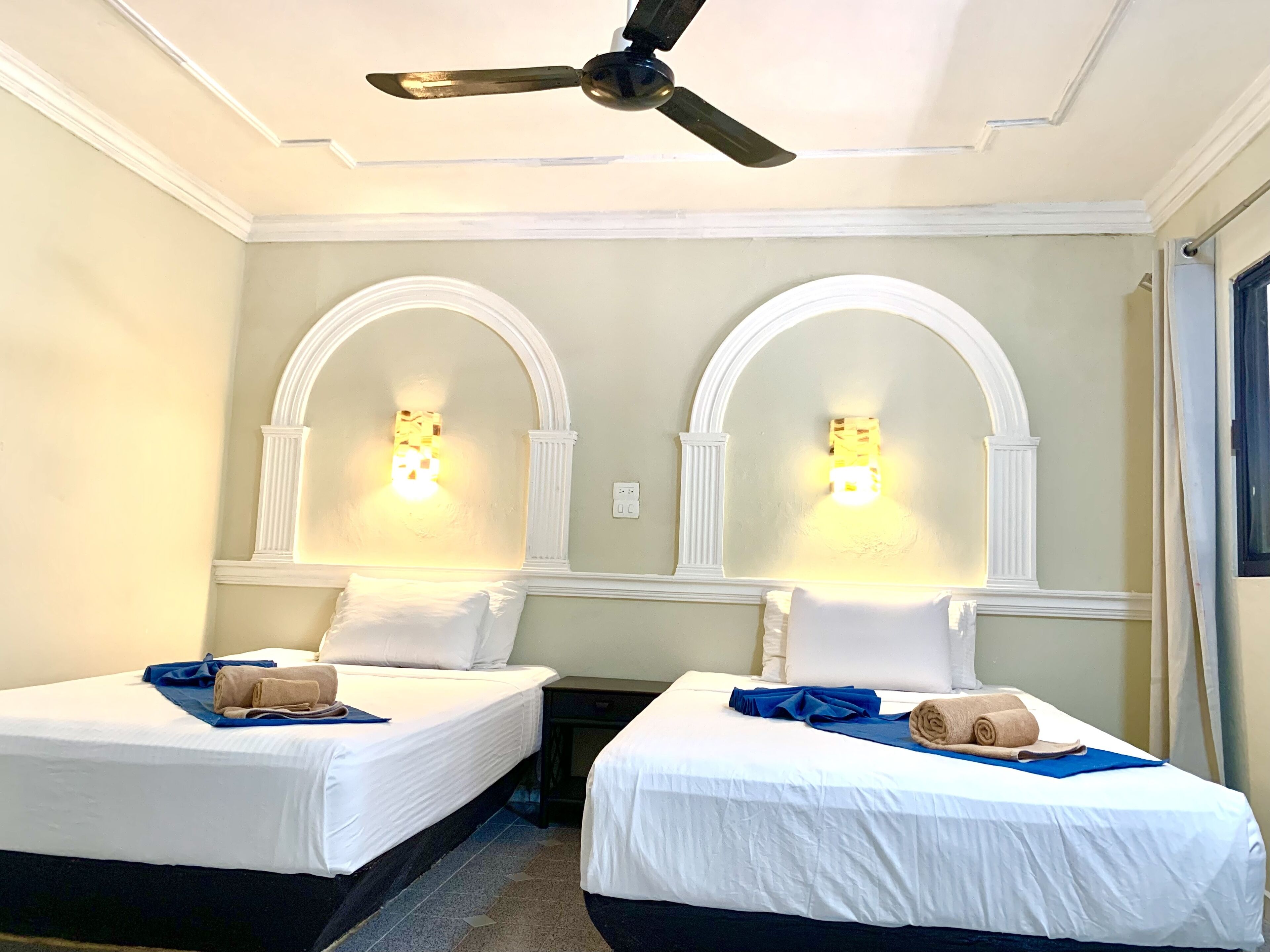 A hotel room with two beds, each with a white bedspread, blue towels, and a ceiling fan.