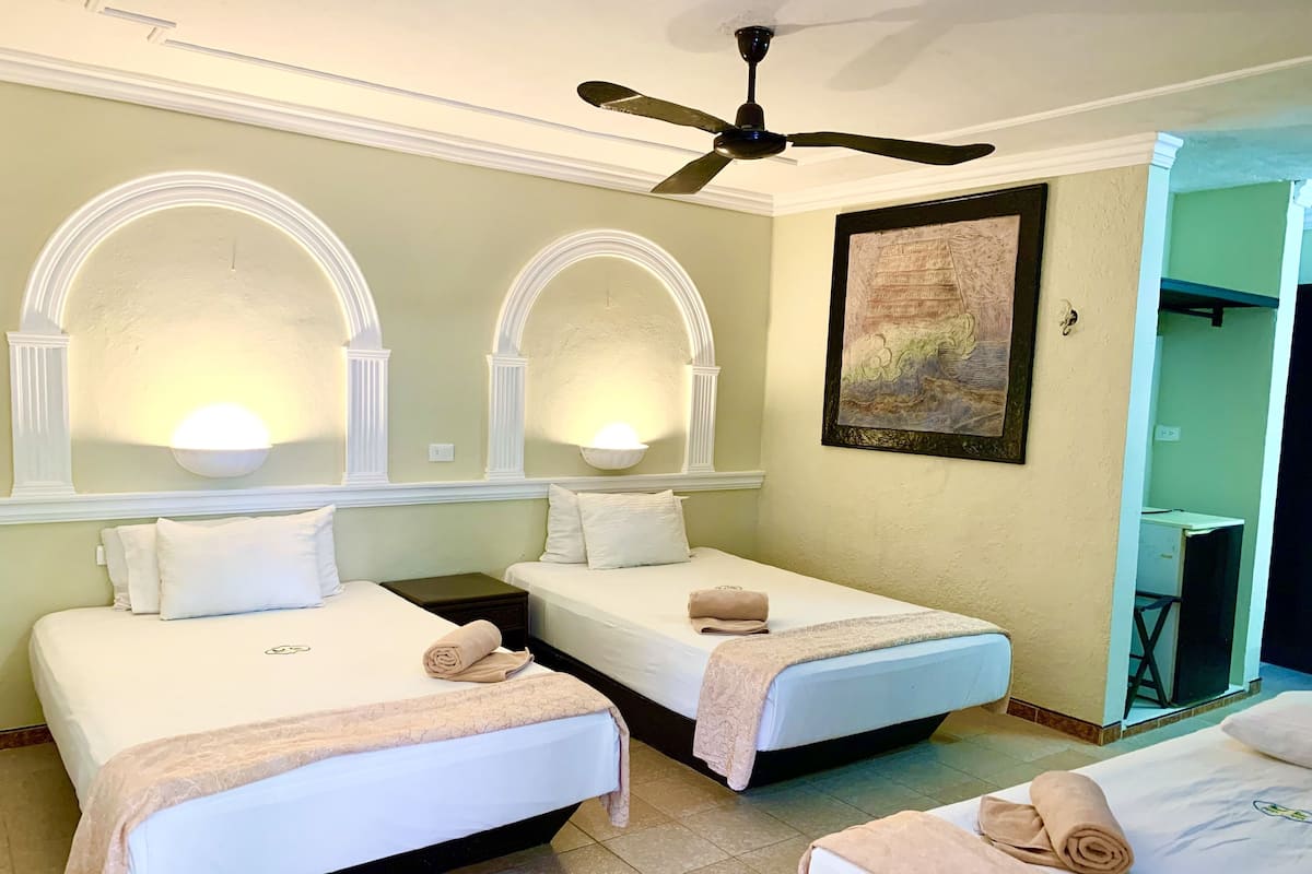 A hotel room with two beds, each with a rolled towel, a ceiling fan, and a painting on the wall.