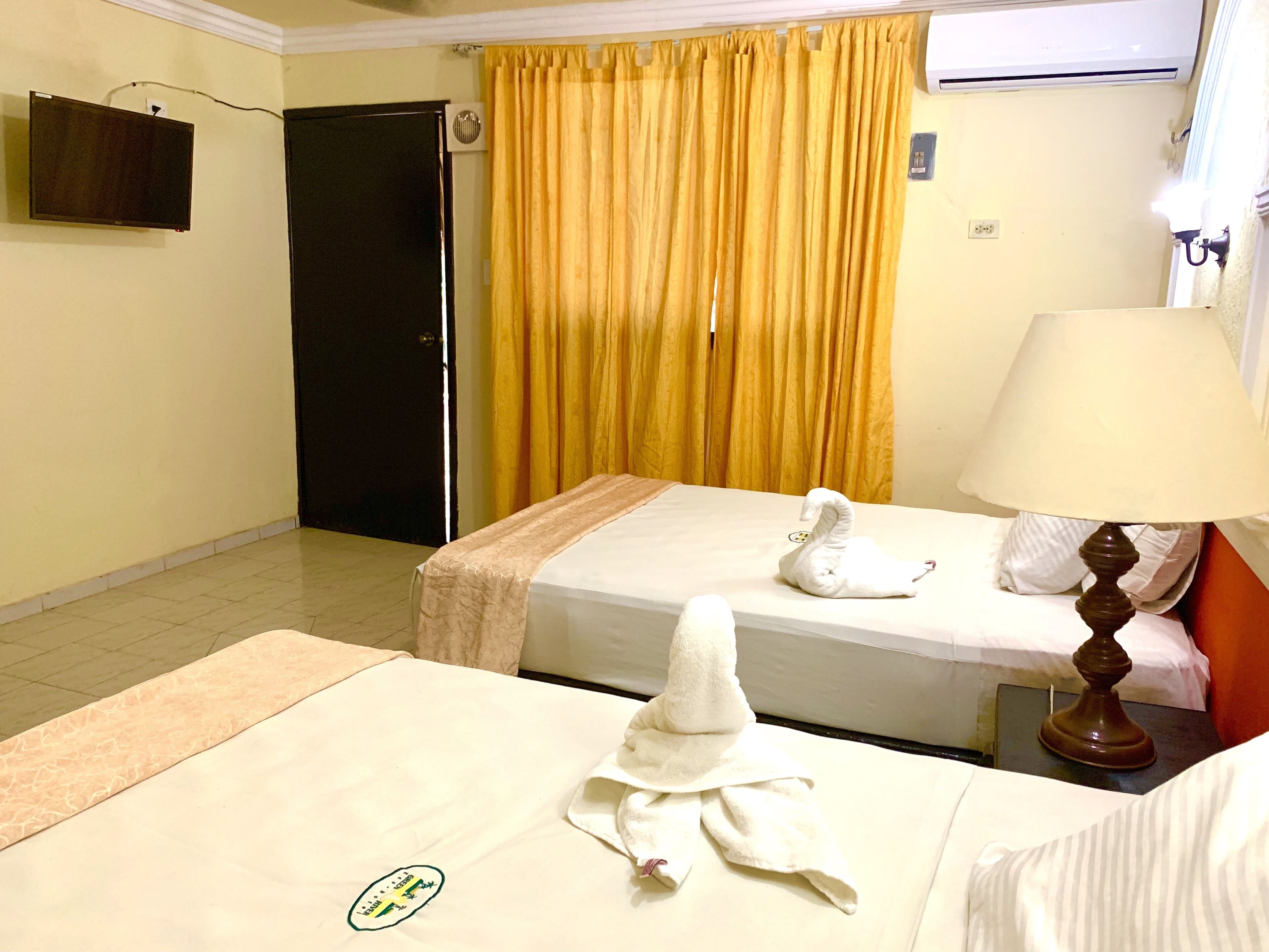 Une chambre d’hôtel avec deux lits, un téléviseur fixé au mur, une porte et une fenêtre avec des rideaux jaunes.
