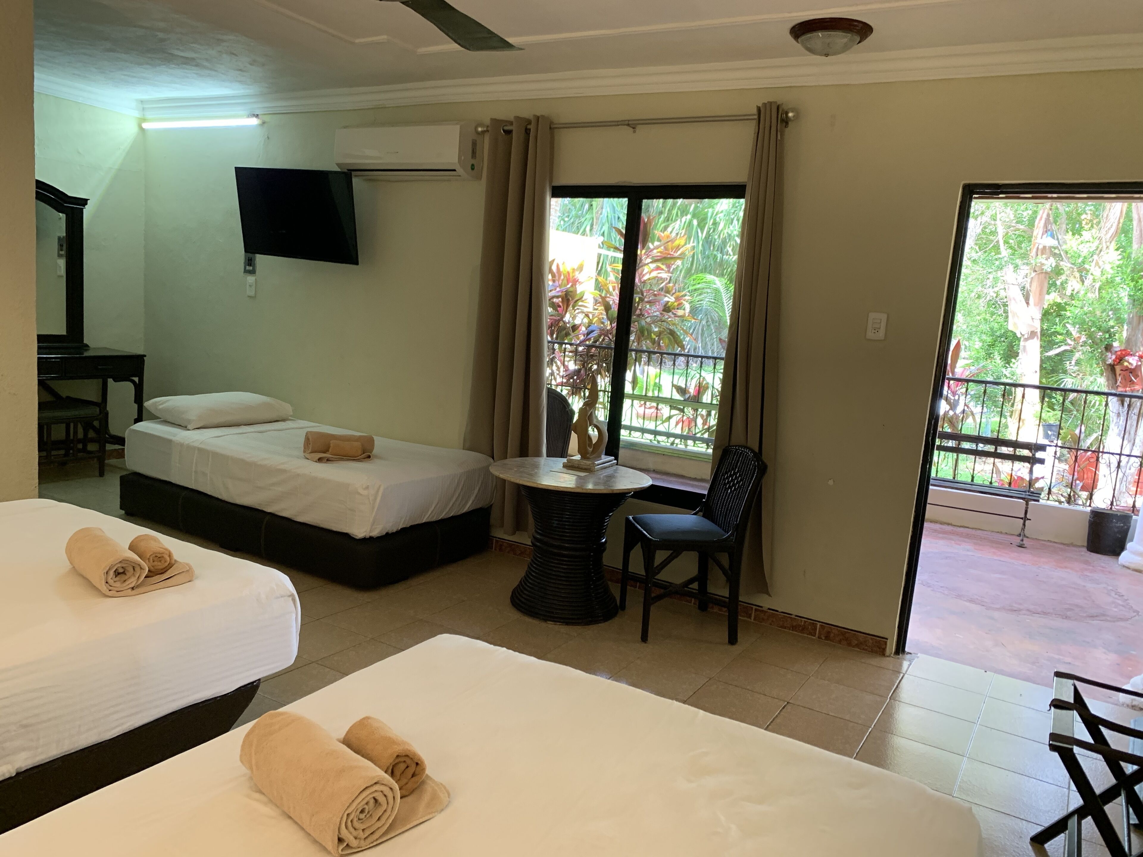 A hotel room with two beds, a desk, a TV, and a balcony with a view of greenery.