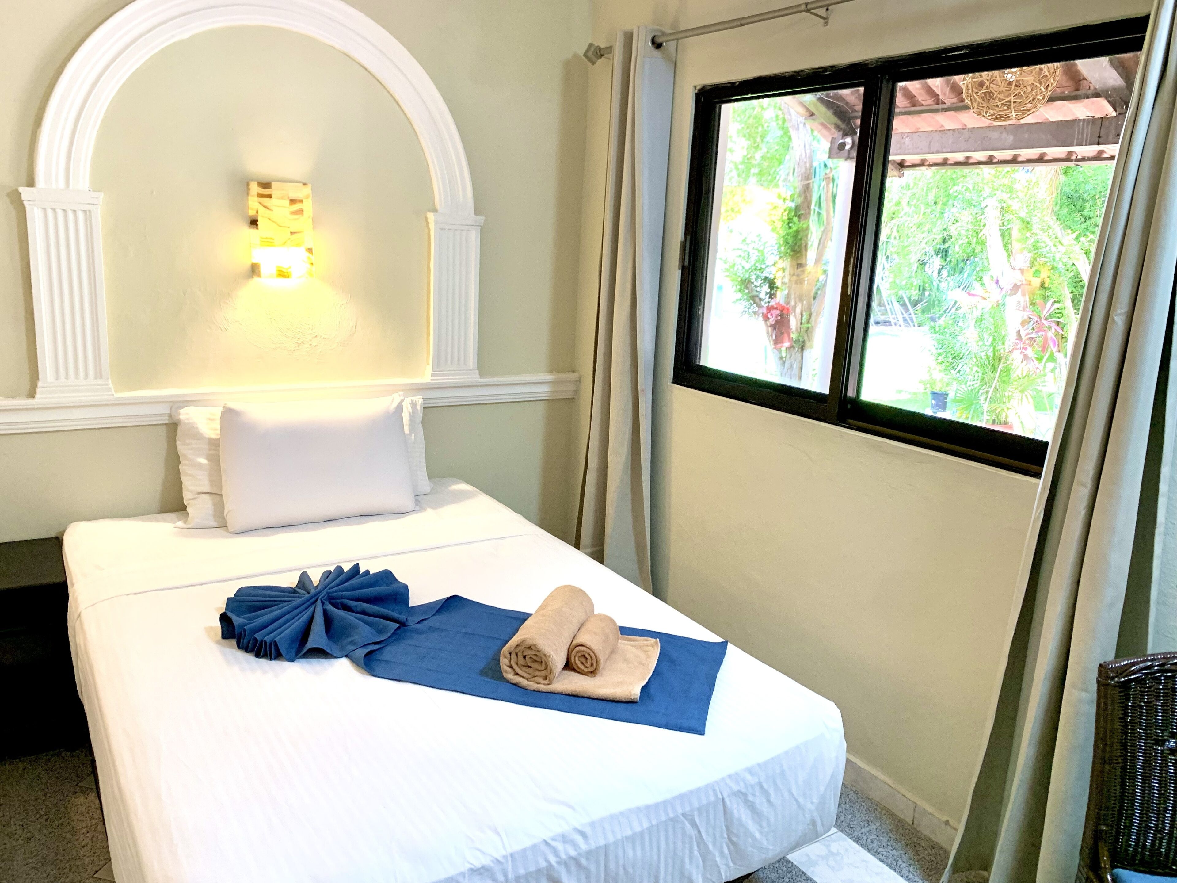 Cama bem arrumada com roupa de cama branca, toalha azul e toalha enrolada. Parede branca com arco decorativo e luminária fixada na parede. Janela com vista para vegetação.