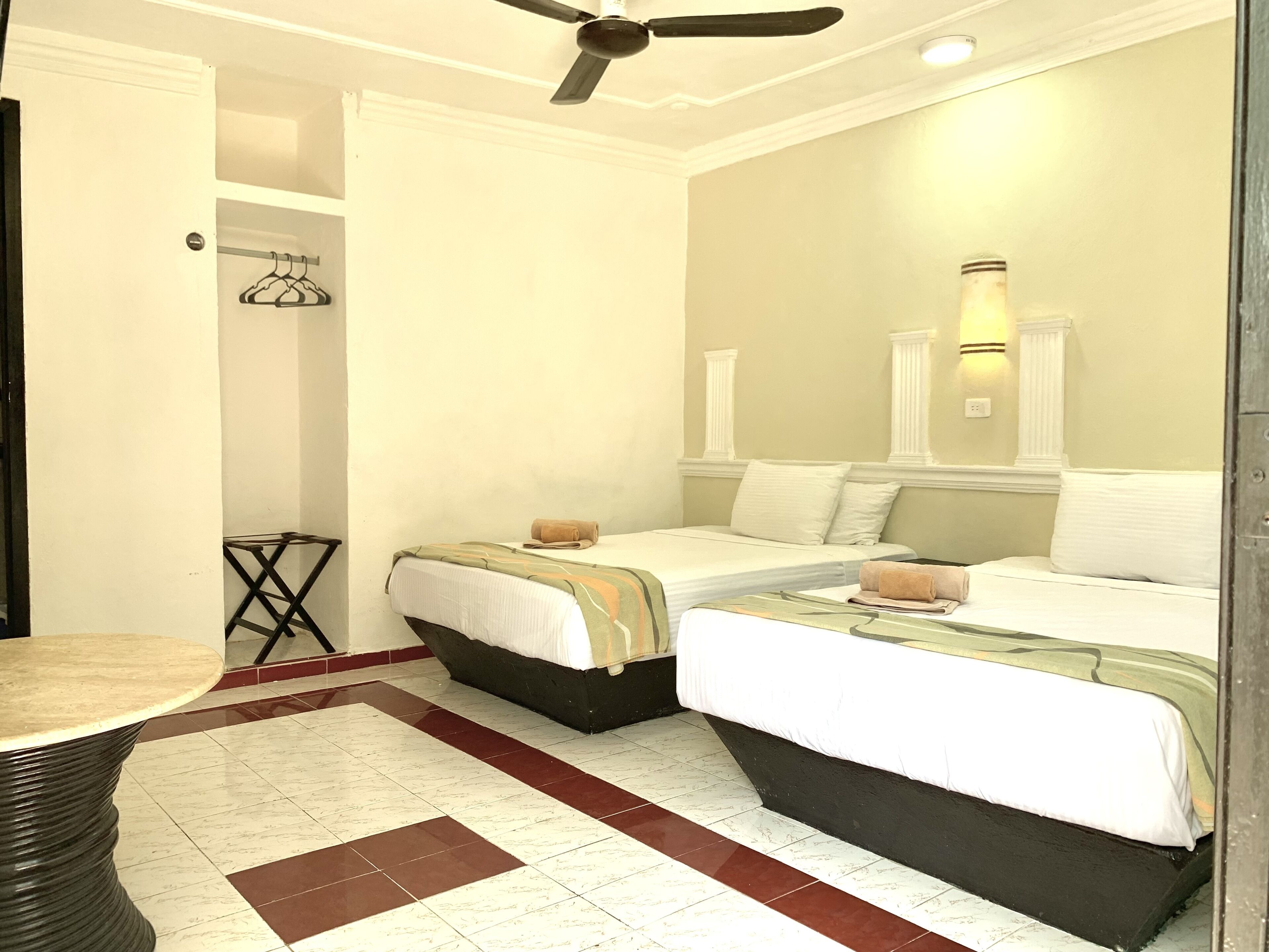 A hotel room with two beds, a ceiling fan, a small table, and a wall-mounted light fixture.
