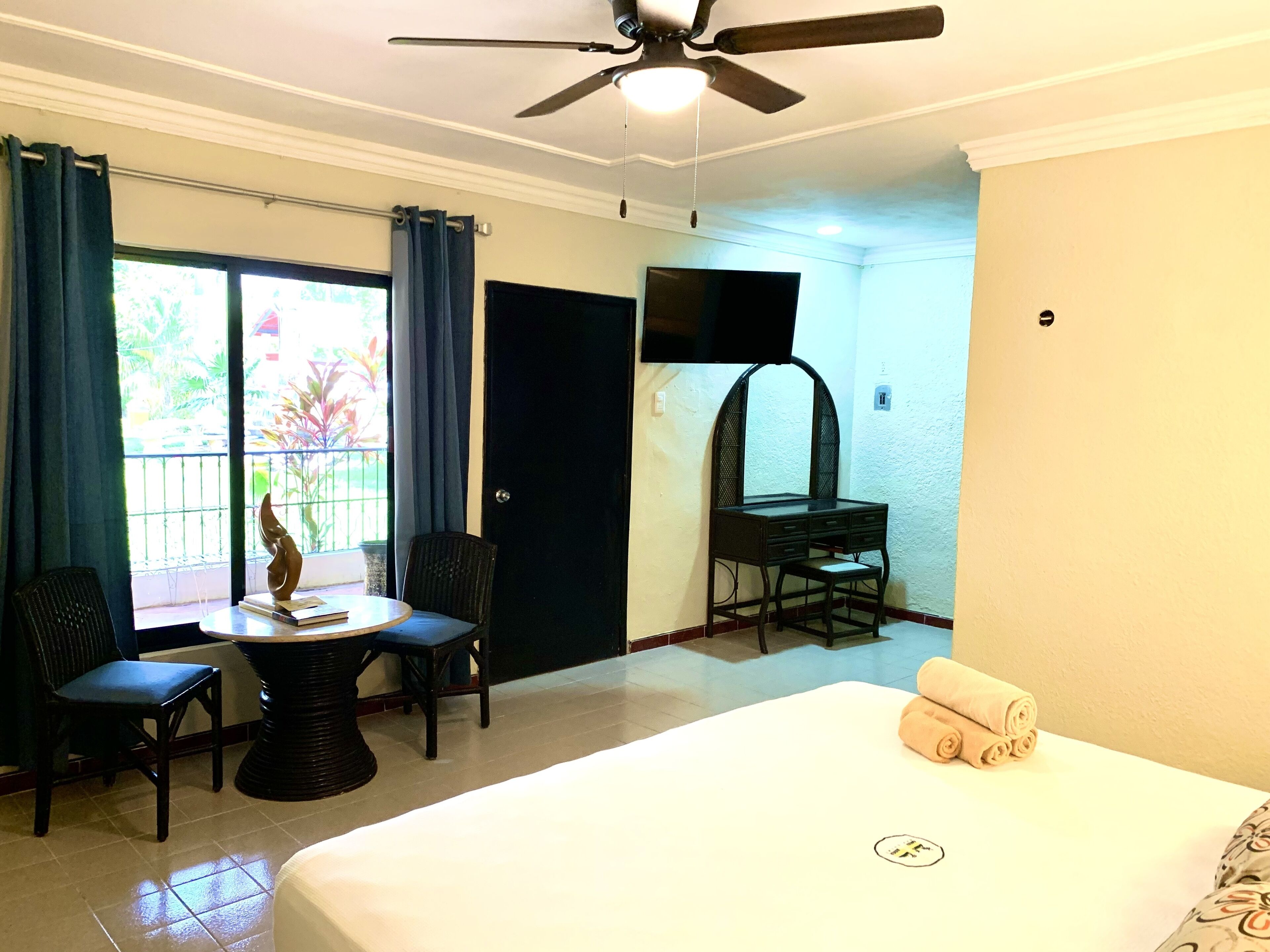 A hotel room with a bed, a desk with a chair, a ceiling fan, a TV mounted on the wall, and a window with curtains.