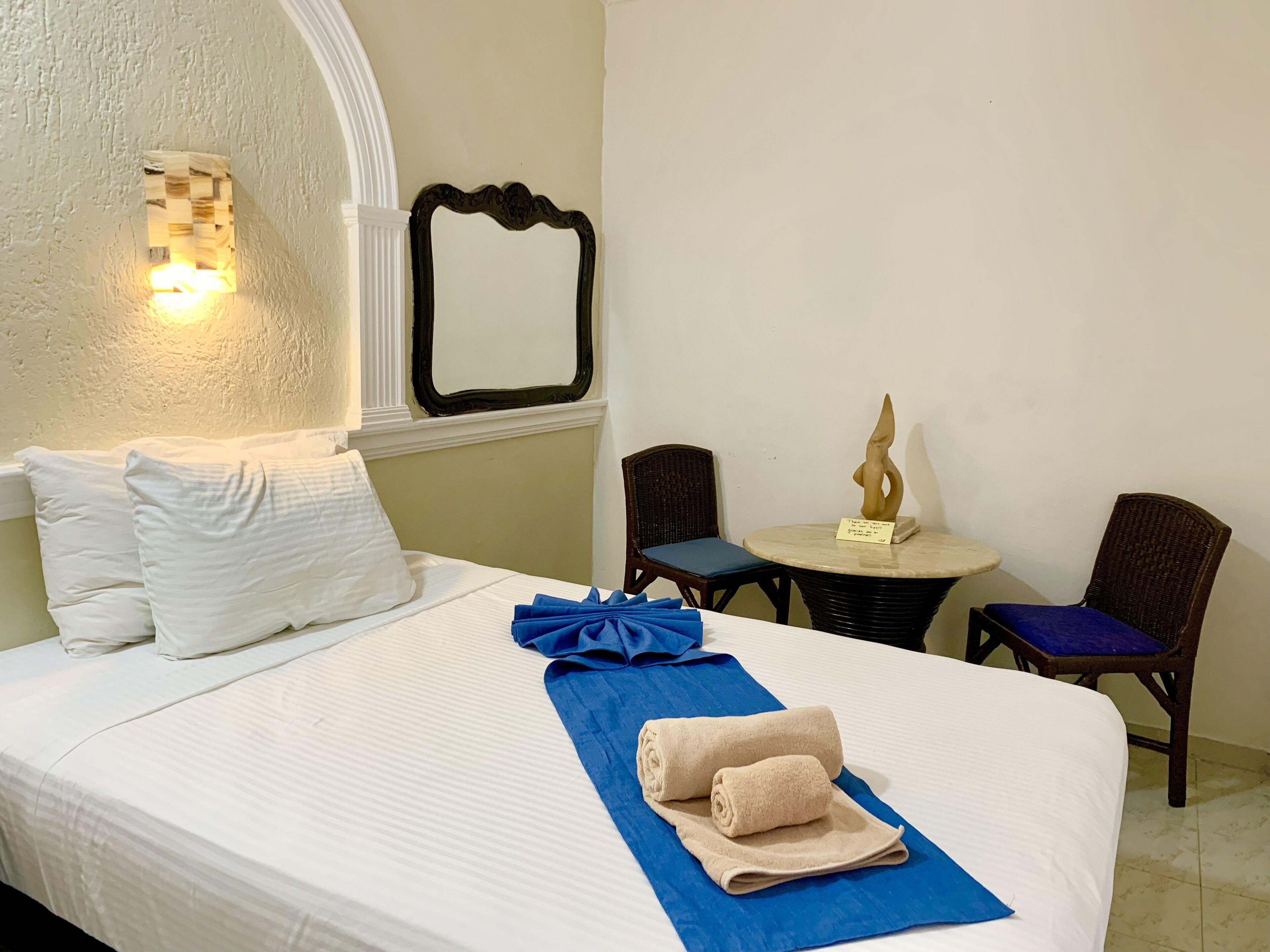 A neatly made bed with white linens, a blue towel, and a folded beige towel. There is a small round table with a statue on it, and two wicker chairs with blue cushions.