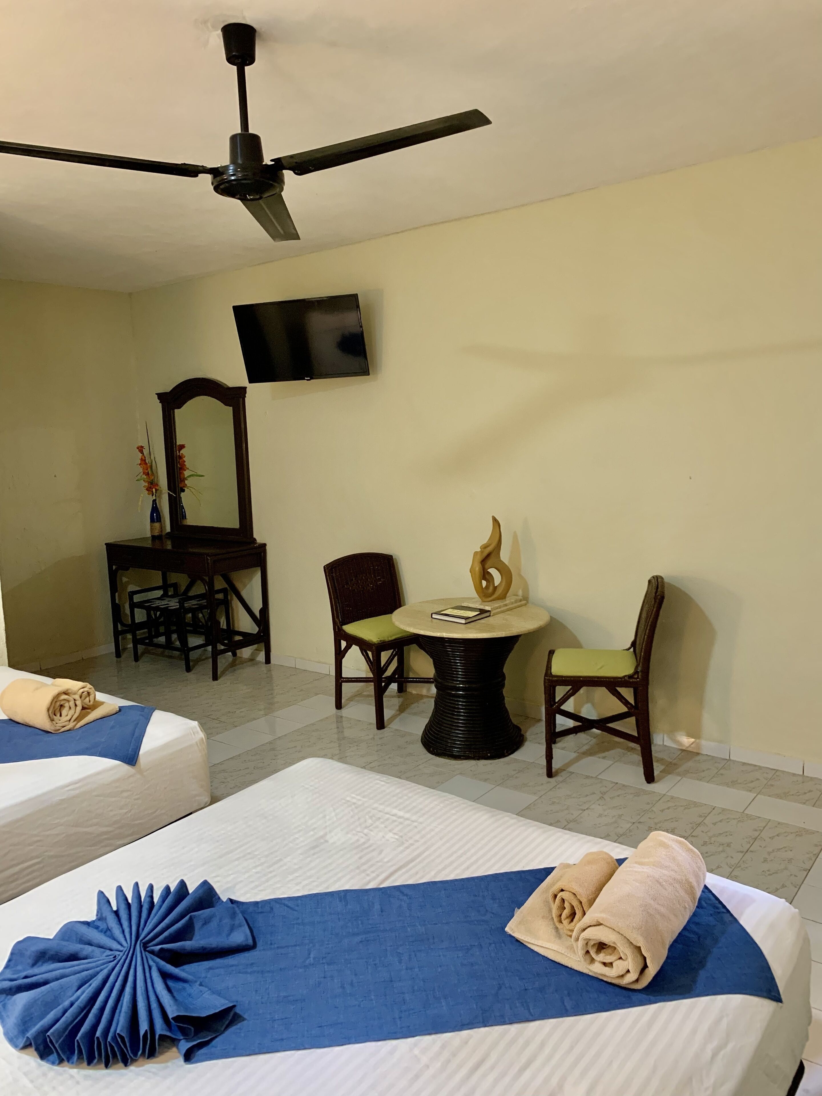 A hotel room with two beds, a ceiling fan, a TV, a small table with a decorative item, and two chairs.
