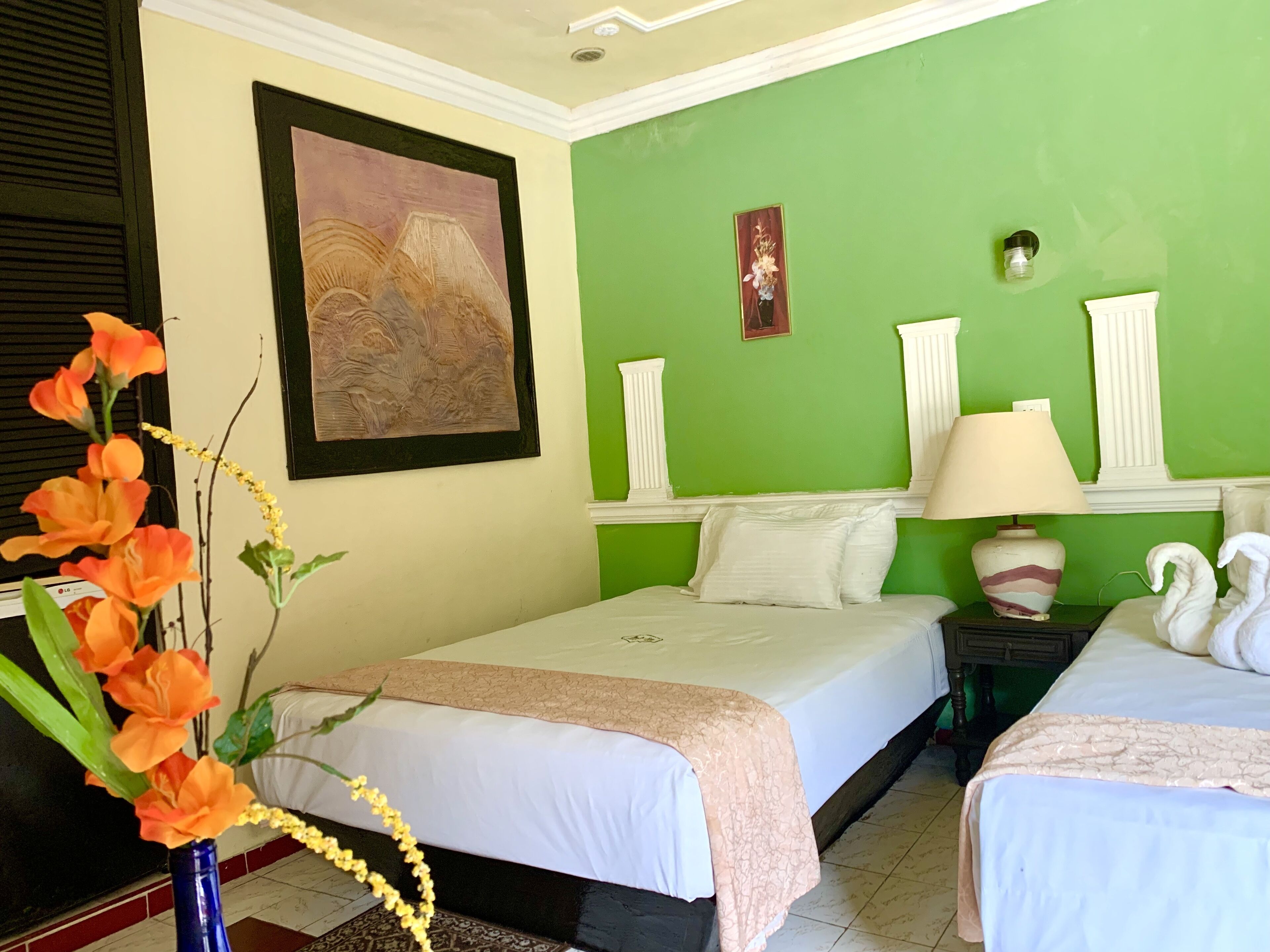 A hotel room with two beds, a green accent wall, a framed painting, a vase with orange flowers, and a lamp.