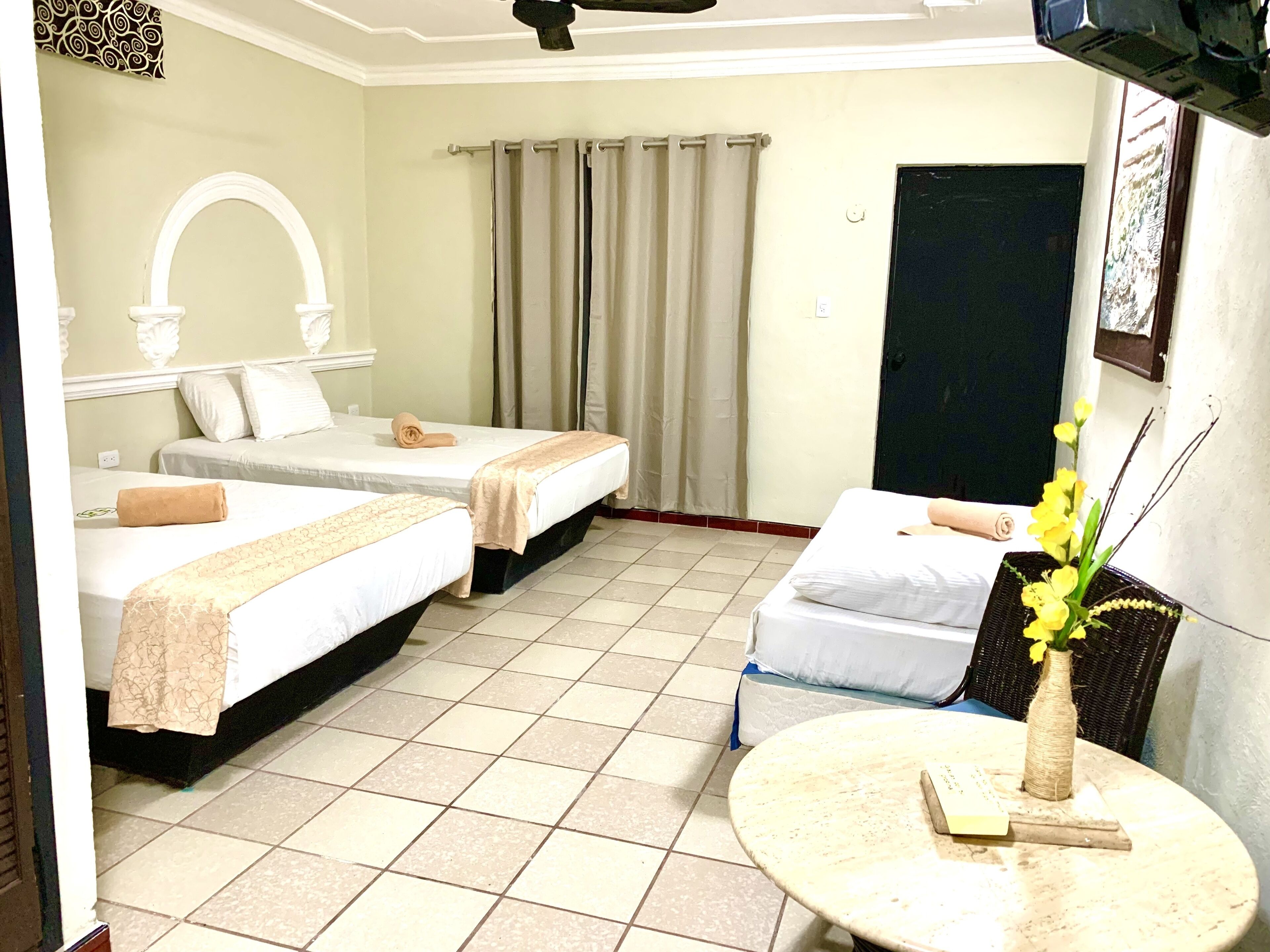 A hotel room with two beds, a small table, a vase with flowers, and a TV mounted on the wall.