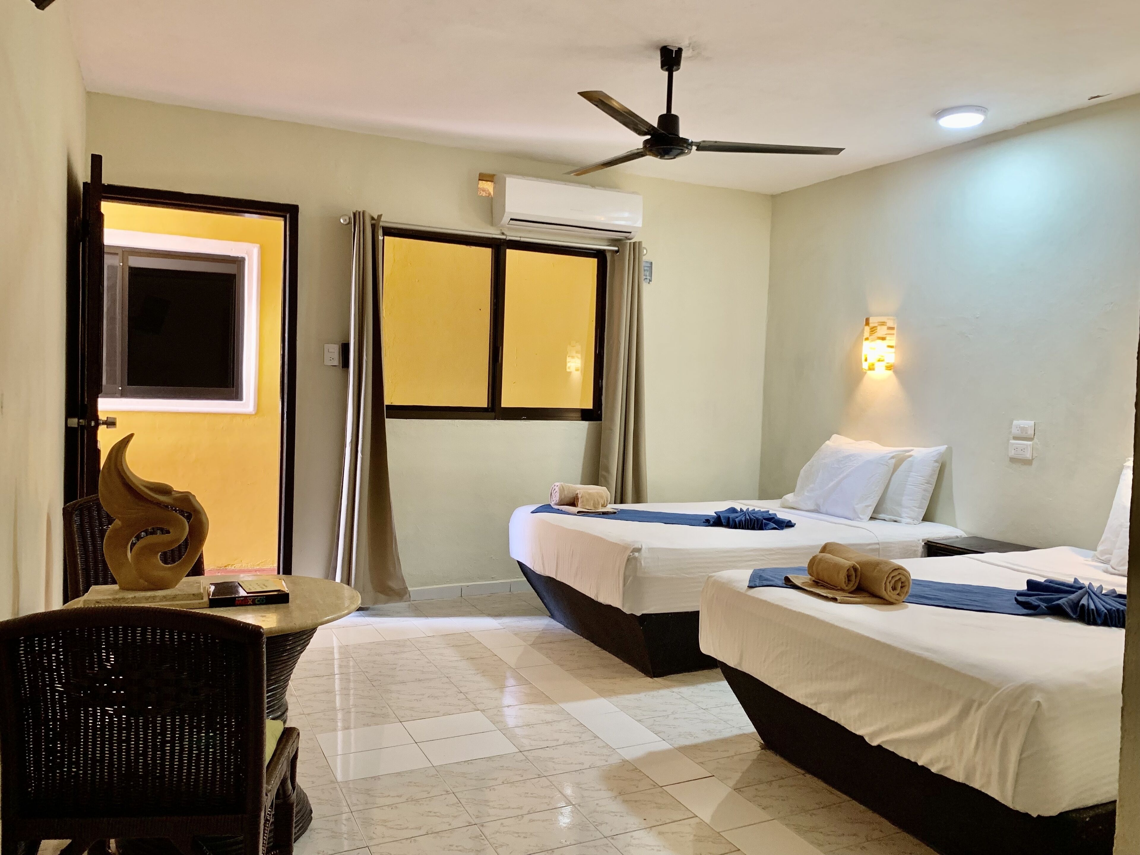 Habitación de hotel con dos camas, ventilador de techo, unidad de aire acondicionado, escritorio con silla y un adorno en la pared.