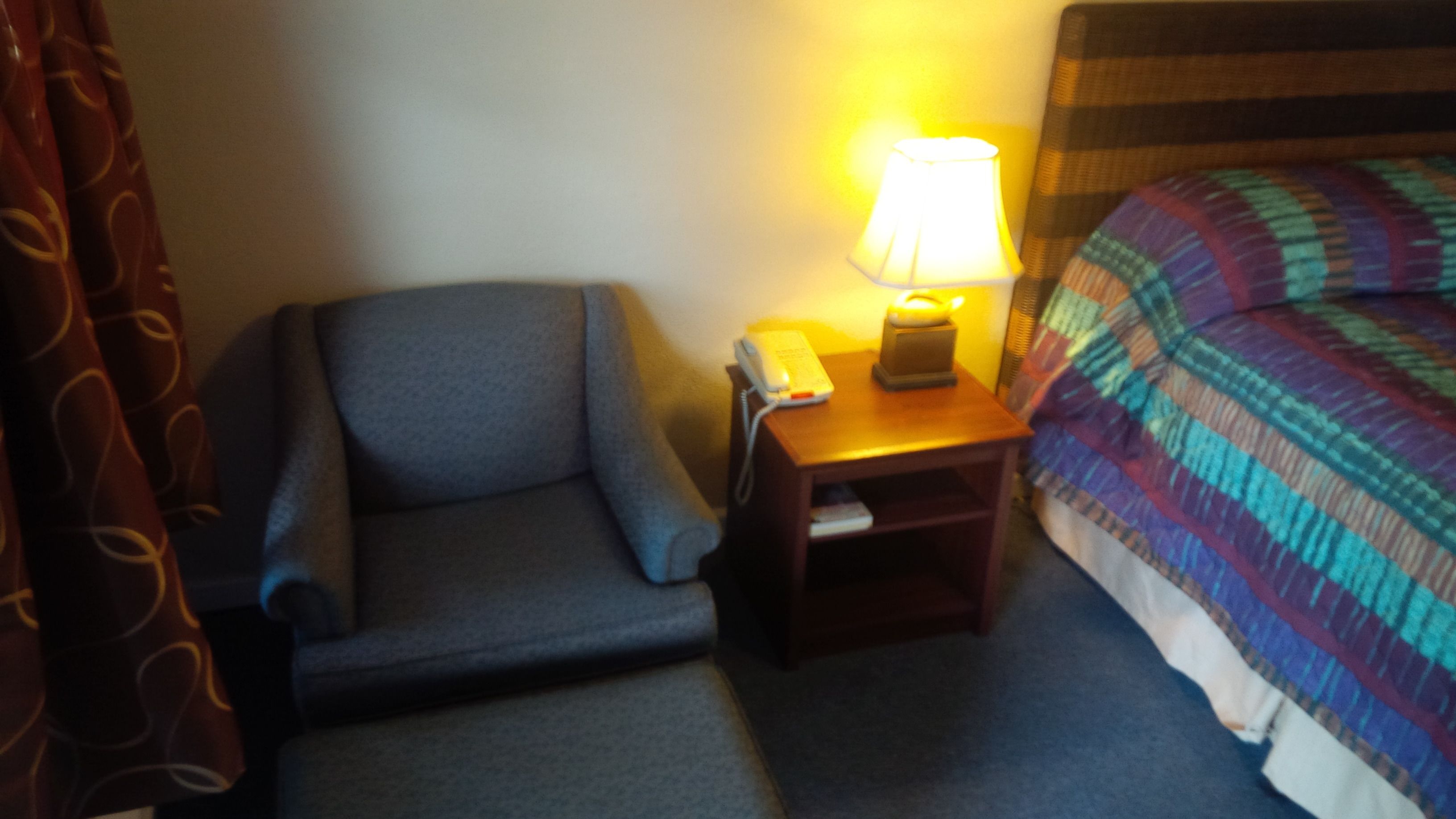 A hotel room with a single bed, a small bedside table with a lamp, a chair, and a curtain.