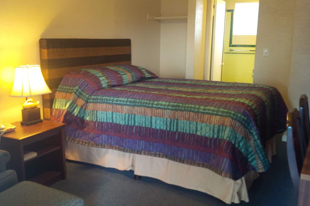 A hotel room with a bed, a bedside table with a lamp, a chair, and a desk with a telephone.