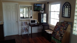 A room with a wooden floor, a white door, a television mounted on the wall, a small desk with a microwave, and a window with blinds. - Holiday Lodge (Port Angeles)