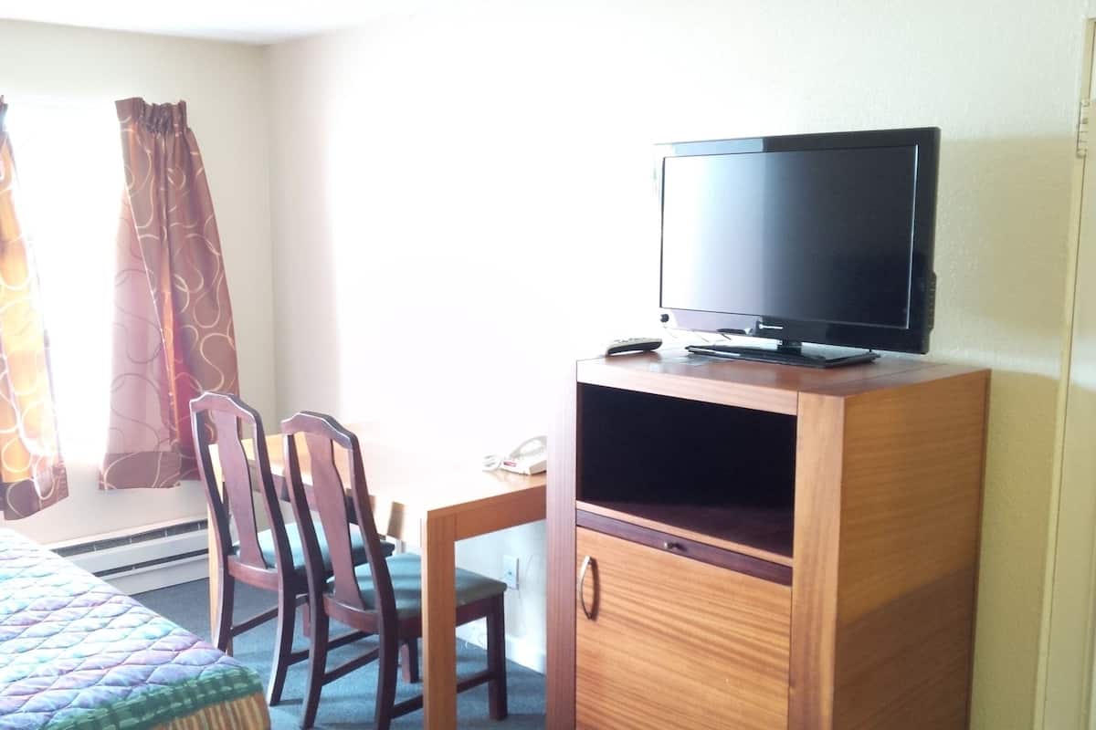 A hotel room with a bed, a wooden desk with a television, two chairs, and a window with curtains.