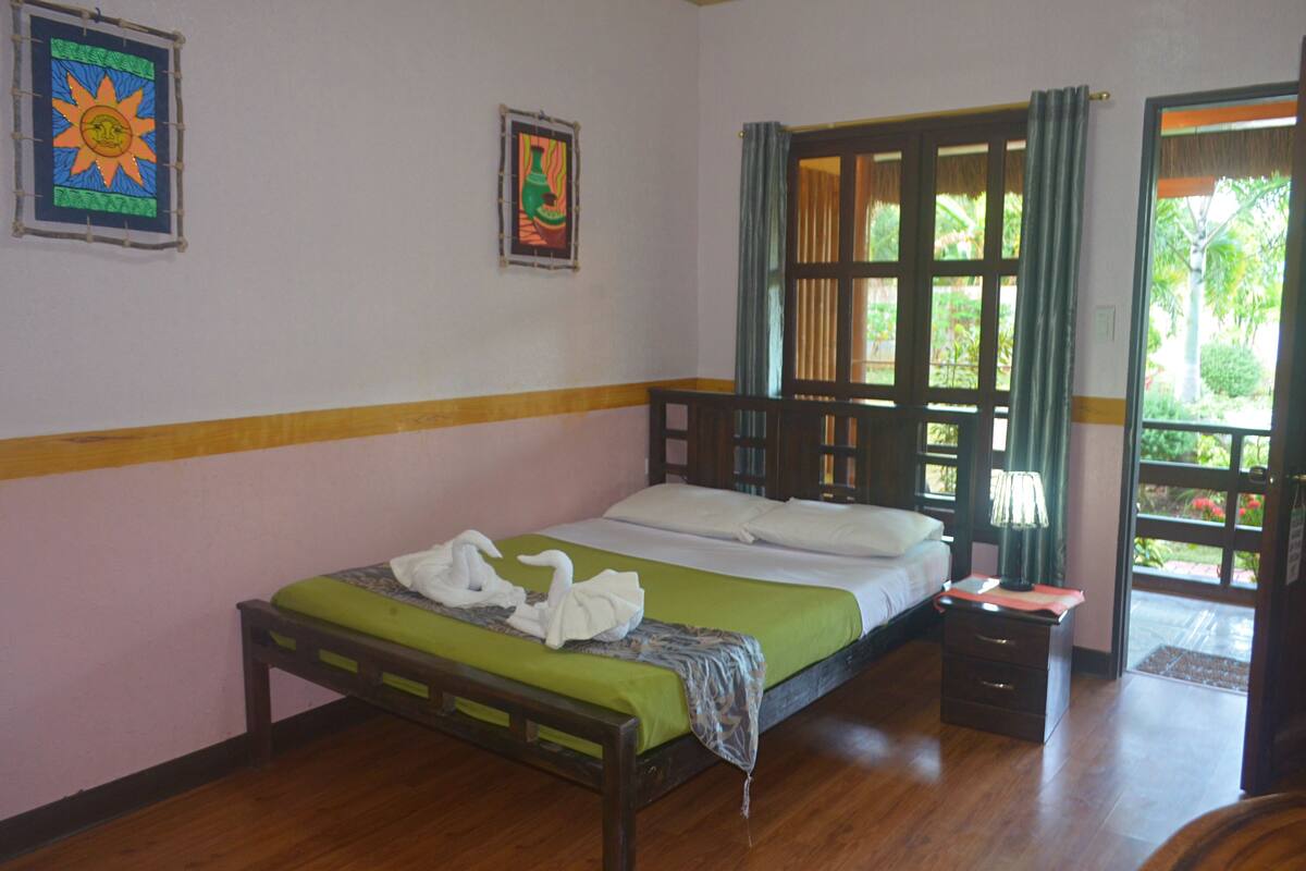 Una cama individual con cubrecama verde y almohadas blancas, cabecera de madera, mesita de noche con lámpara y ventana con cortinas.