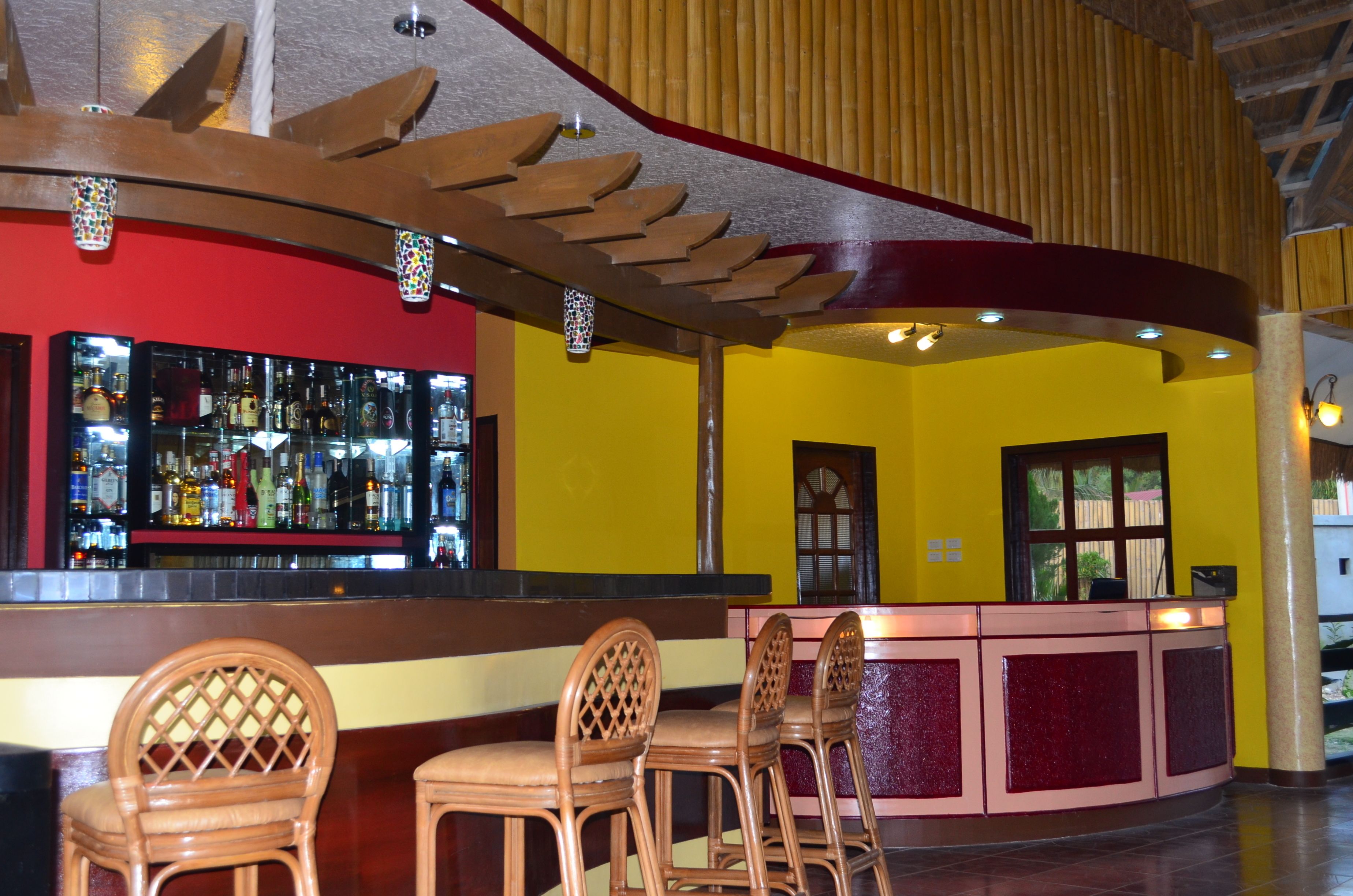 A bar area with a wooden ceiling, a red wall, and a yellow wall. There are three wooden chairs and a bar counter with bottles.