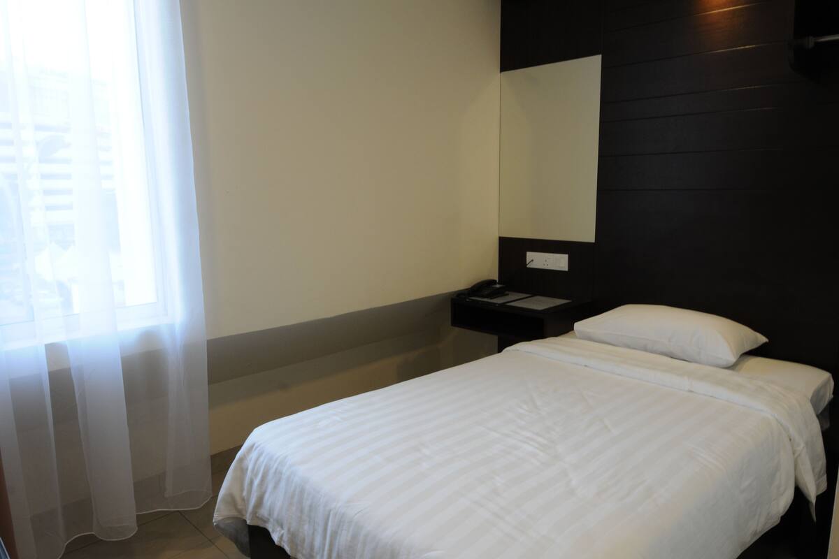 A hotel room with a bed, a nightstand, a mirror, and a window with sheer curtains.