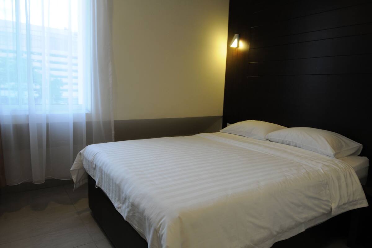 A neatly made bed with white linens in a hotel room with a dark headboard and a window with sheer curtains.