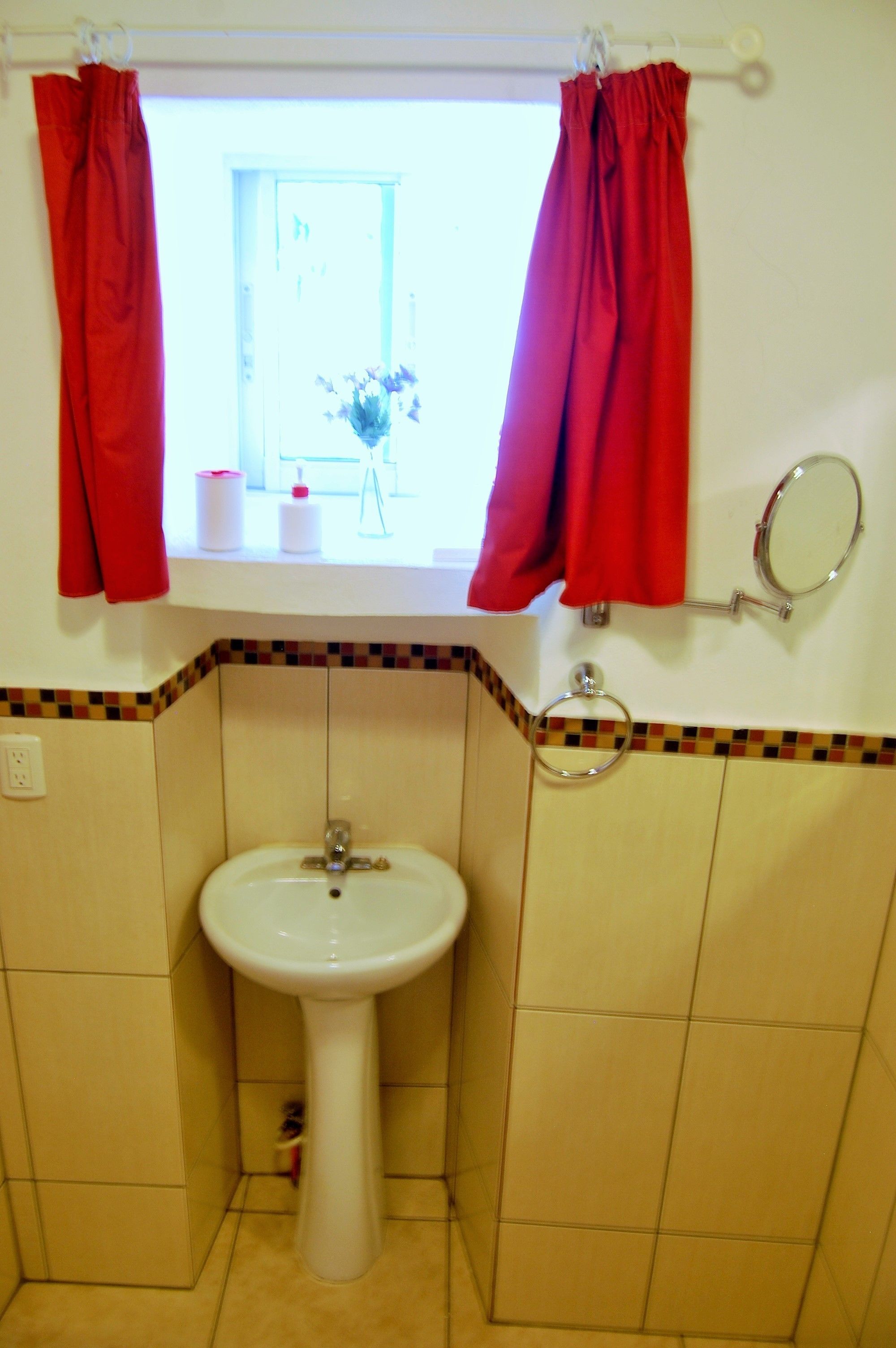 Un baño con un lavamanos blanco, cortinas rojas, un espejo y una ventana que da a una habitación con un jarrón de flores.