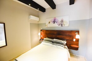 A bedroom with a wooden headboard, a bed with striped bedding, two bedside tables with lamps, an air conditioning unit, and a floral artwork on the wall. - Island Life Hostel (Santo Domingo)