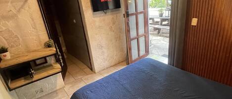 Ein Schlafzimmer mit einem Bett, einem an der Wand montierten Fernseher, einer Holztür und einem kleinen Tisch mit einer Pflanze.