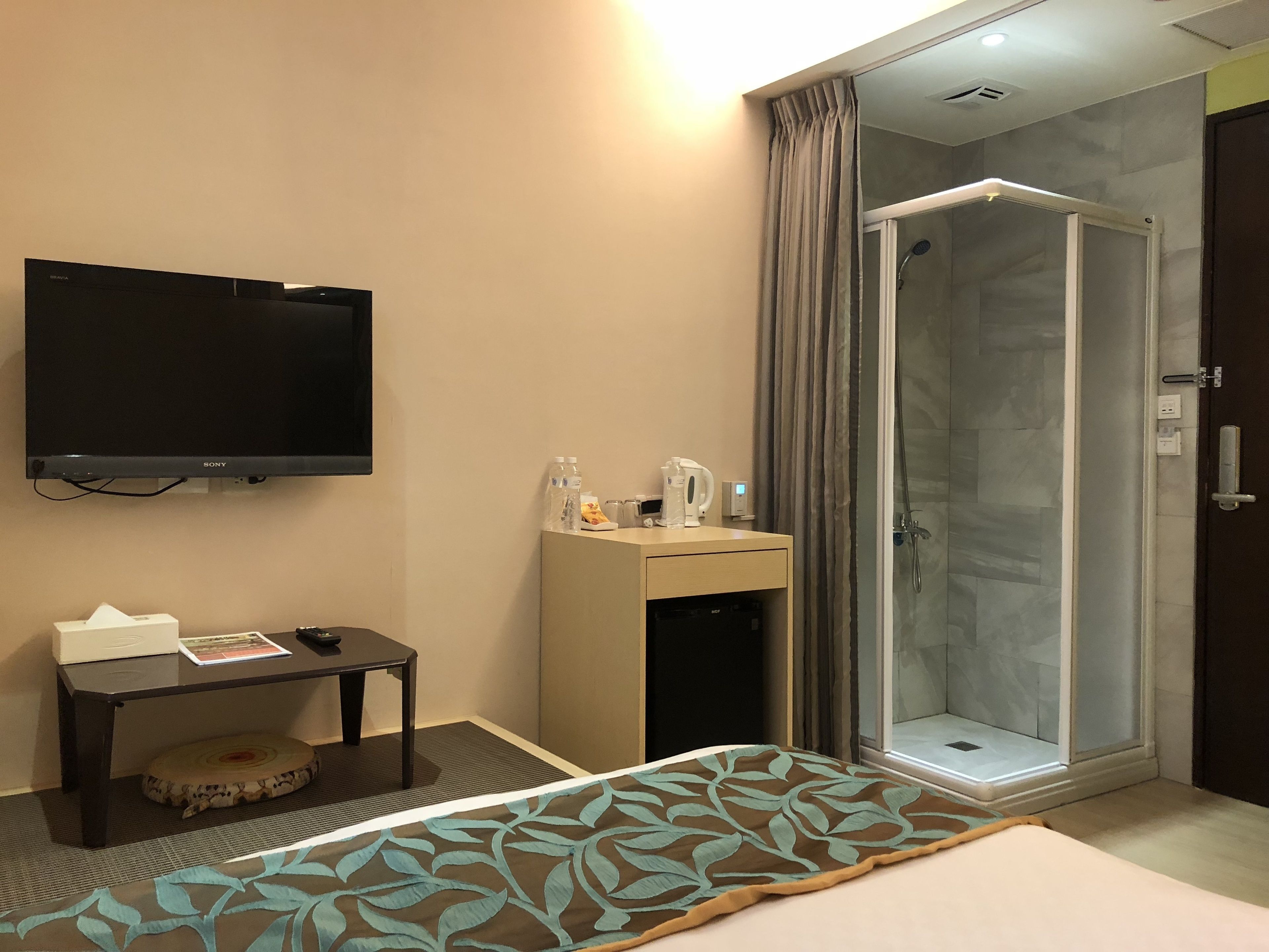 A hotel room with a bed, a TV, a desk with a lamp, a shower, and a small table with a tissue box and a magazine.