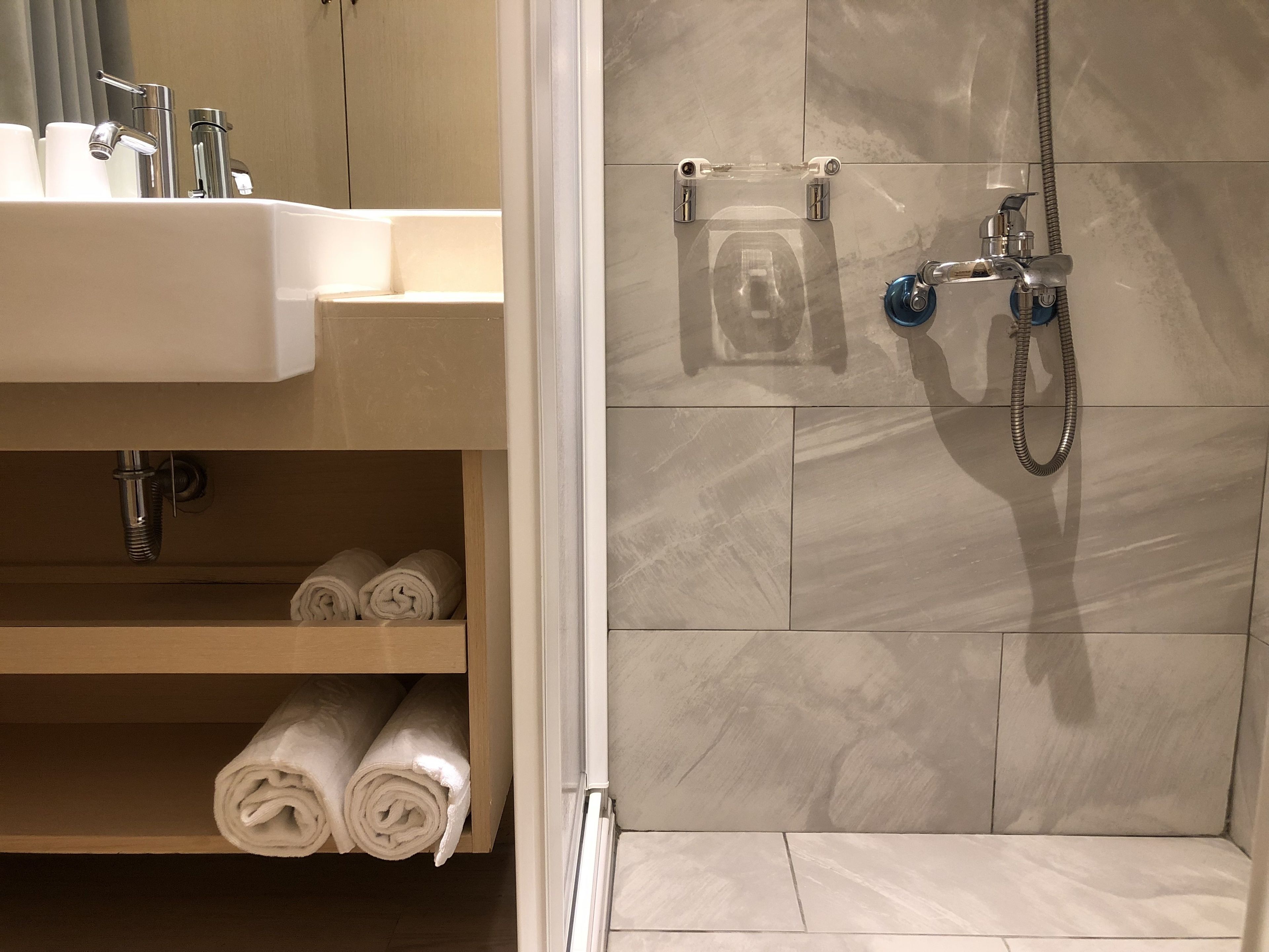 Een badkamer met een wastafel, spiegel en douche. Handdoeken liggen op een plank.