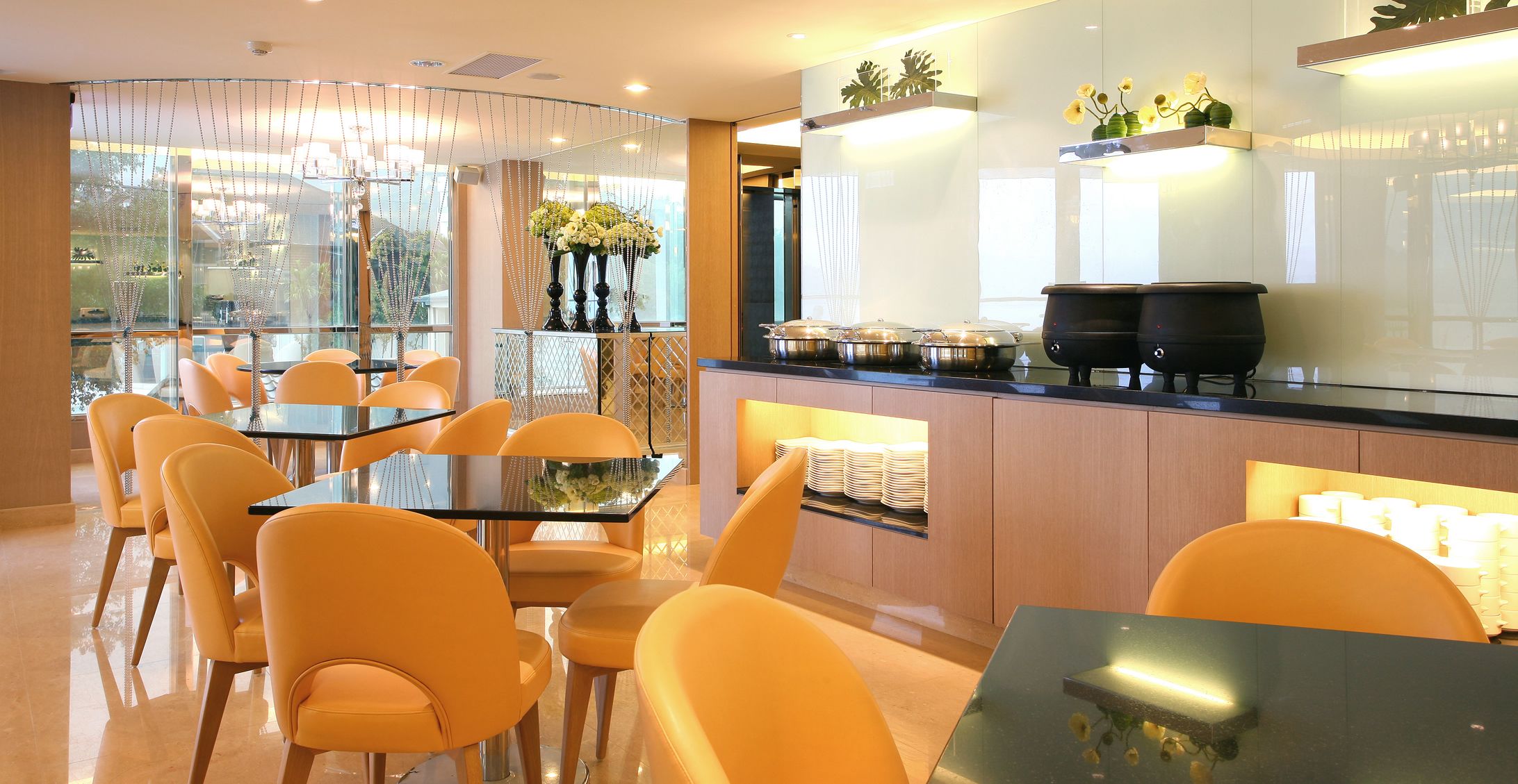 A modern dining area with glass tables and chairs, a kitchen counter with pots and a coffee machine, and a view through large windows.
