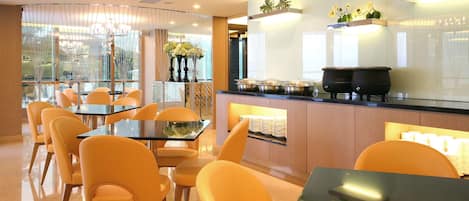 A modern dining area with glass tables and chairs, a kitchen counter with pots and a coffee machine, and a view through large windows.