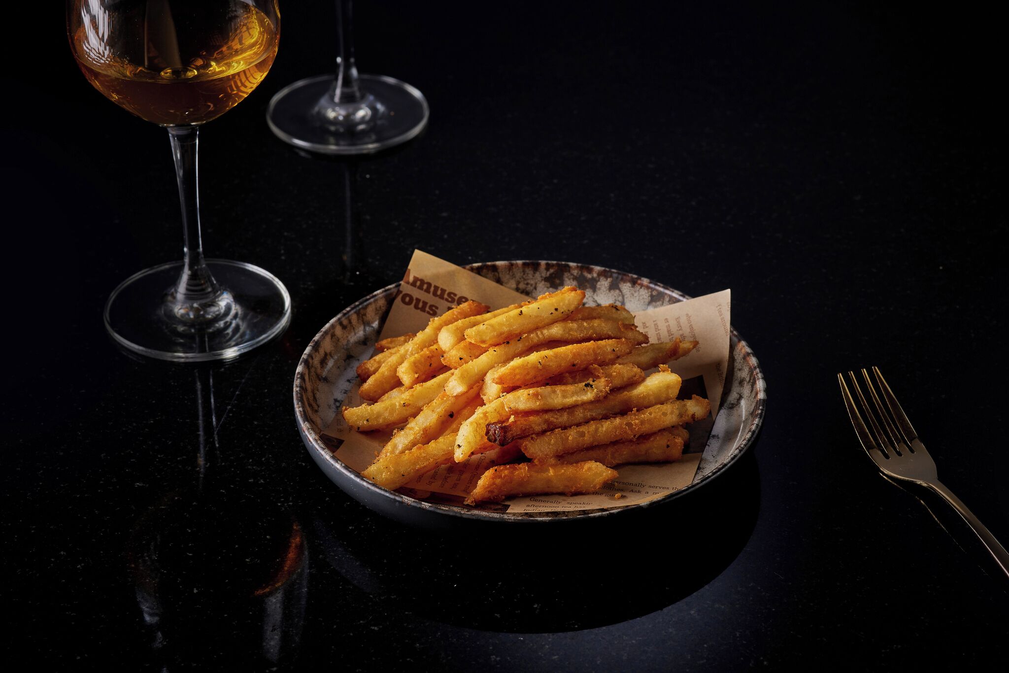 A plate of golden-brown fries with a paper label, a wine glass, and a fork on a dark surface.