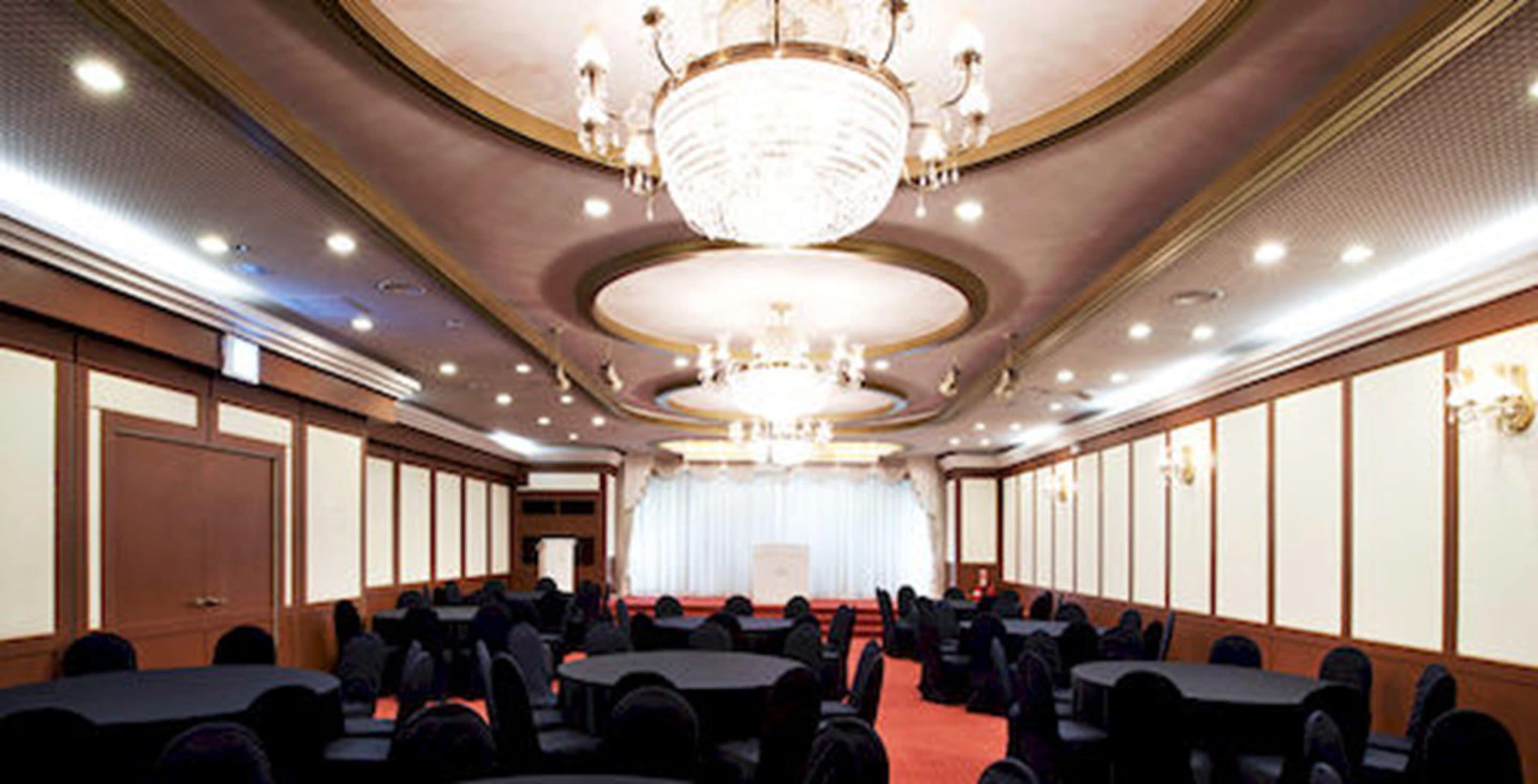 A conference room with round tables and chairs, a chandelier, and a stage at the front.