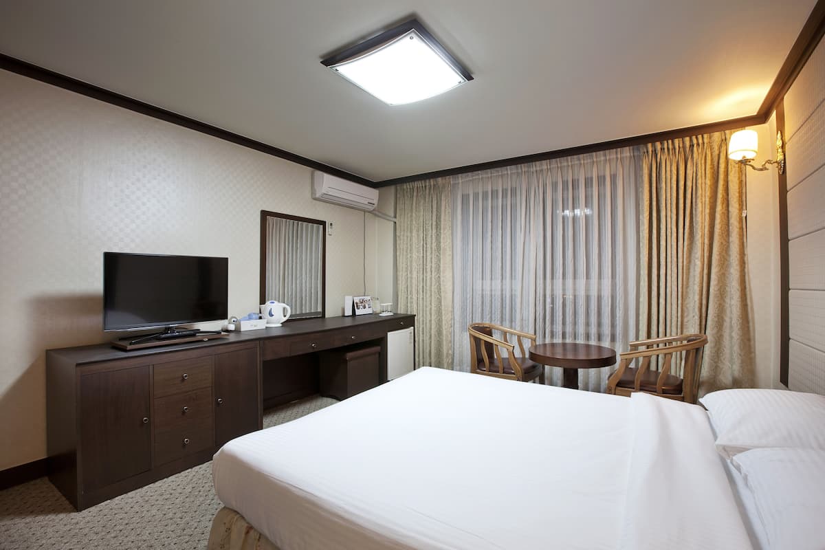 A hotel room with a bed, a desk with a TV, a chair, a small table, and a window with curtains.