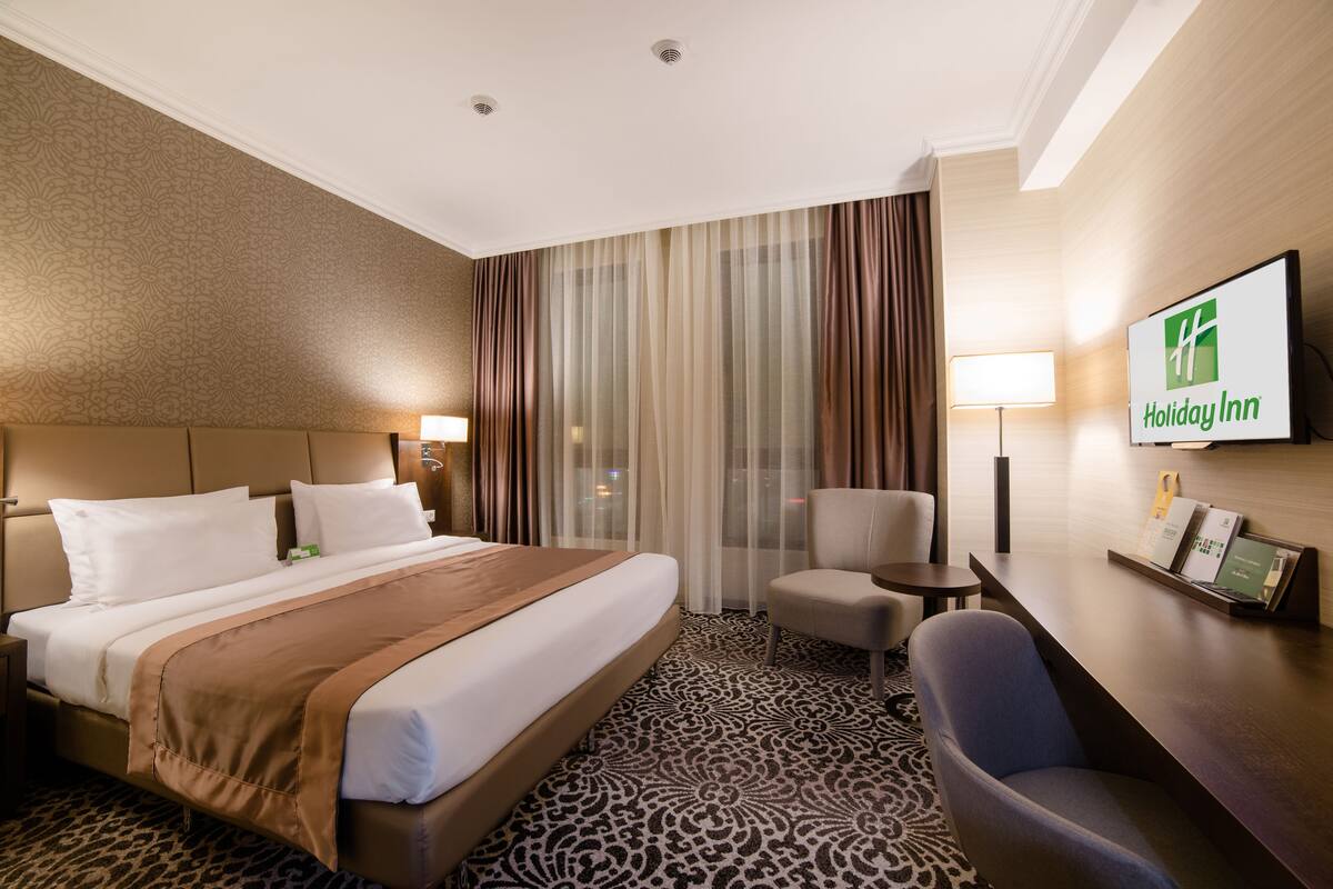 A hotel room with a large bed, a desk, a chair, and a TV displaying the Holiday Inn logo.