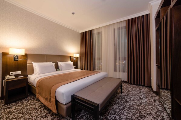 A hotel room with a large bed, a bedside table with a lamp, a bench, and a view of the city through sheer curtains. - Holiday Inn Aktau by IHG (Aktau)