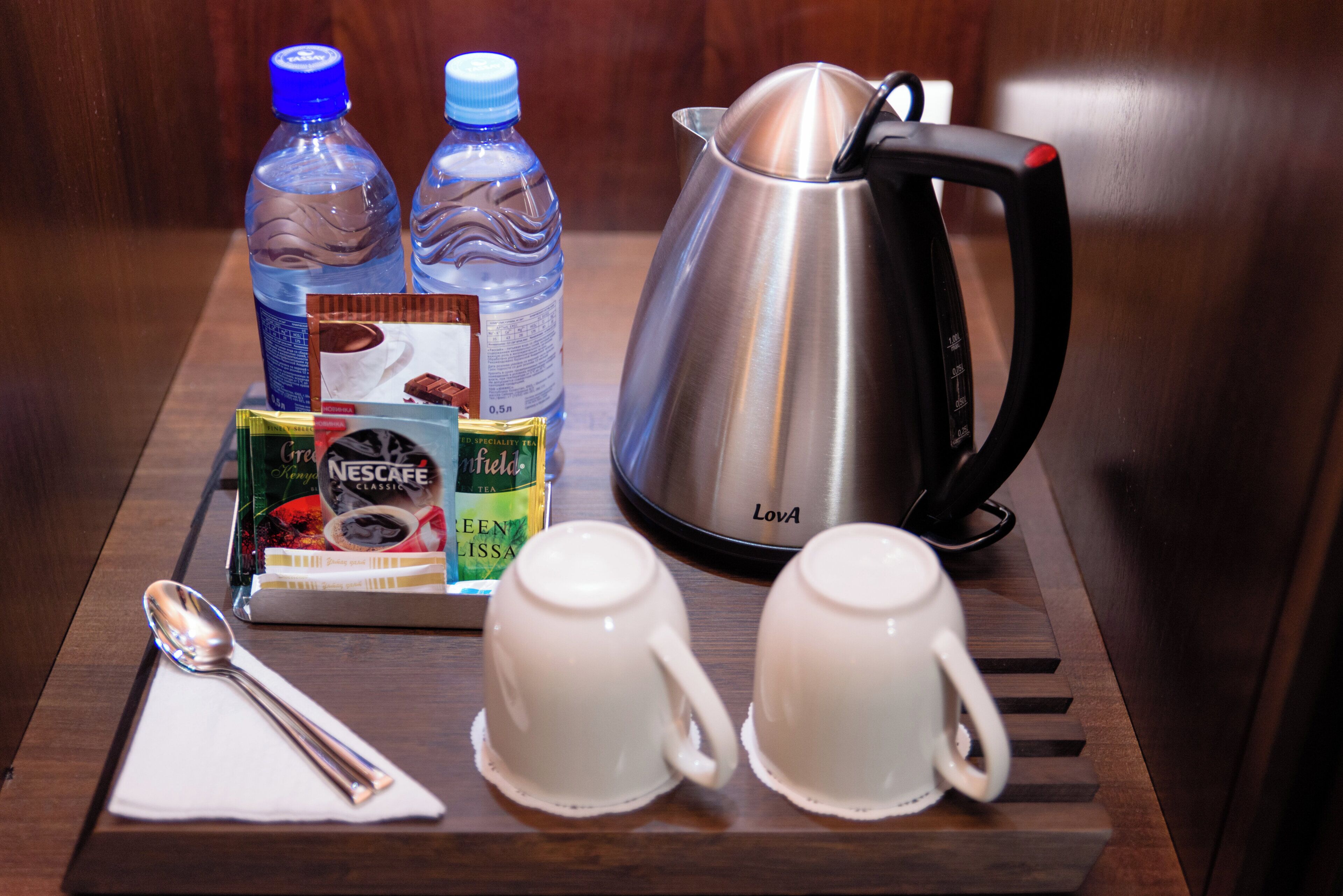 Una bandeja con una cafetera, dos botellas de agua, una cuchara, una servilleta y dos tazas sobre una superficie de madera.