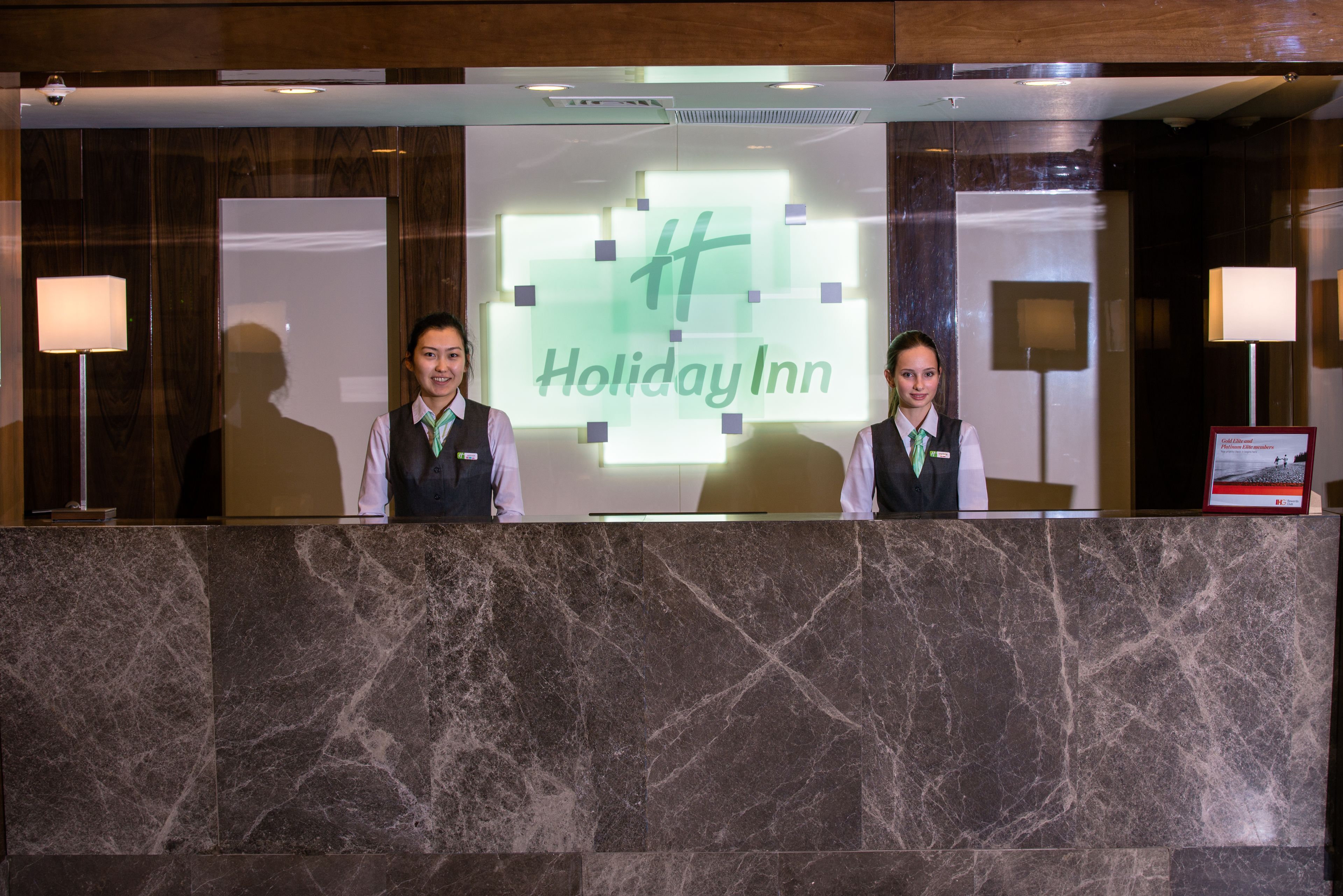 Un mostrador de recepción en un Holiday Inn, con dos recepcionistas, suelo de mármol y un cartel iluminado de gran tamaño.