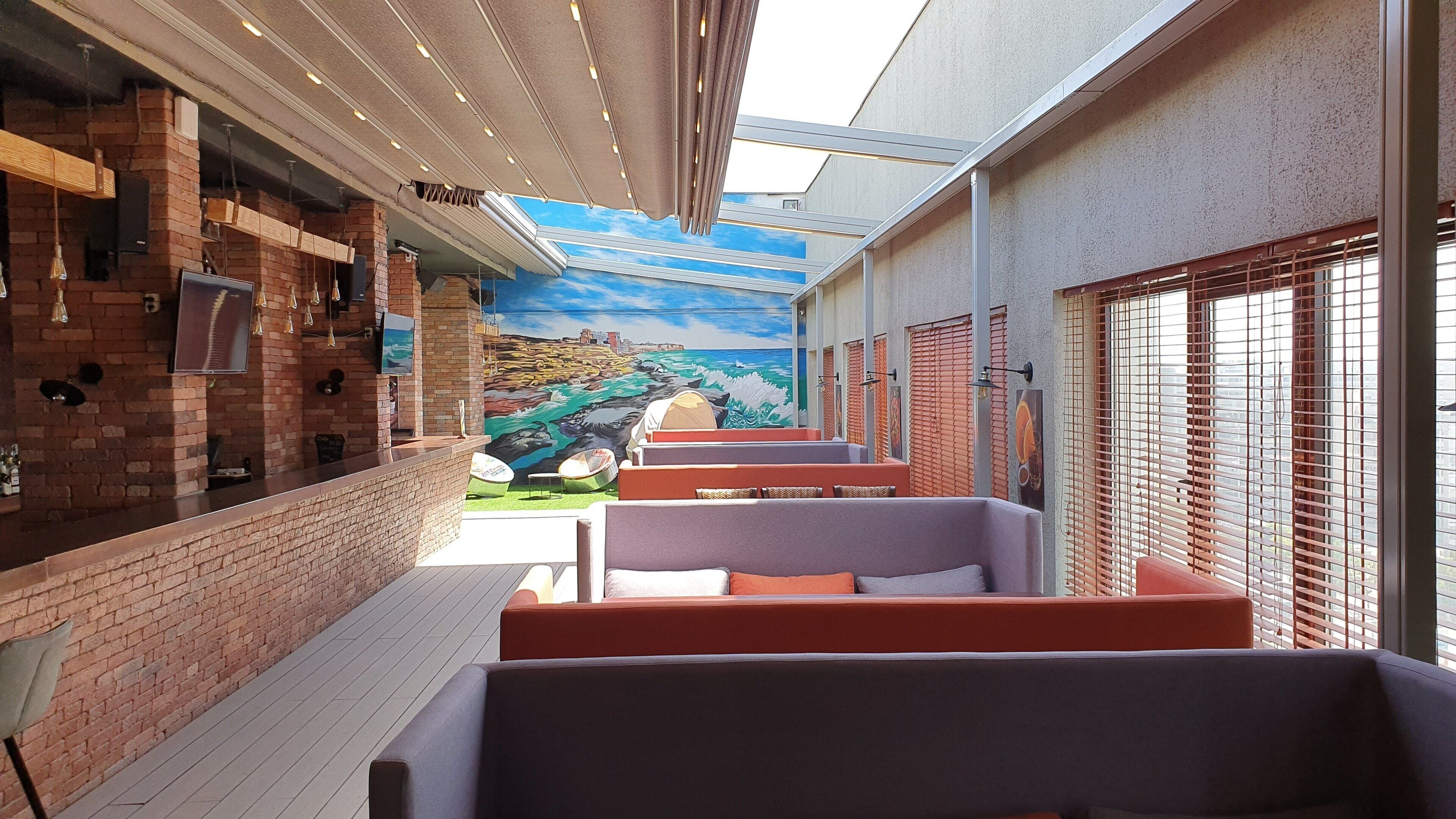 A covered outdoor seating area with brick walls, a mural of a coastal scene, and a view of the city.