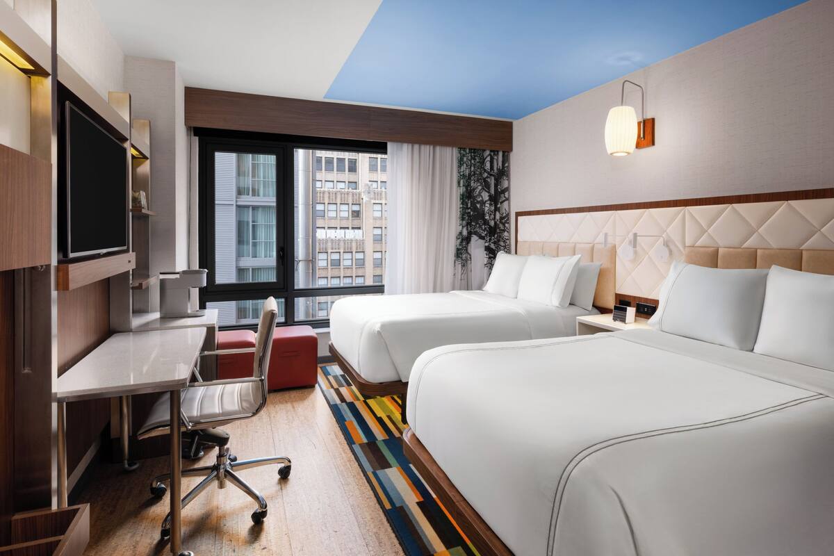 A hotel room with two beds, a desk, a TV, and a window with a city view.