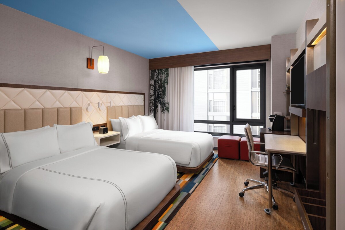 A hotel room with two beds, a desk, a chair, a TV, and a window with curtains.