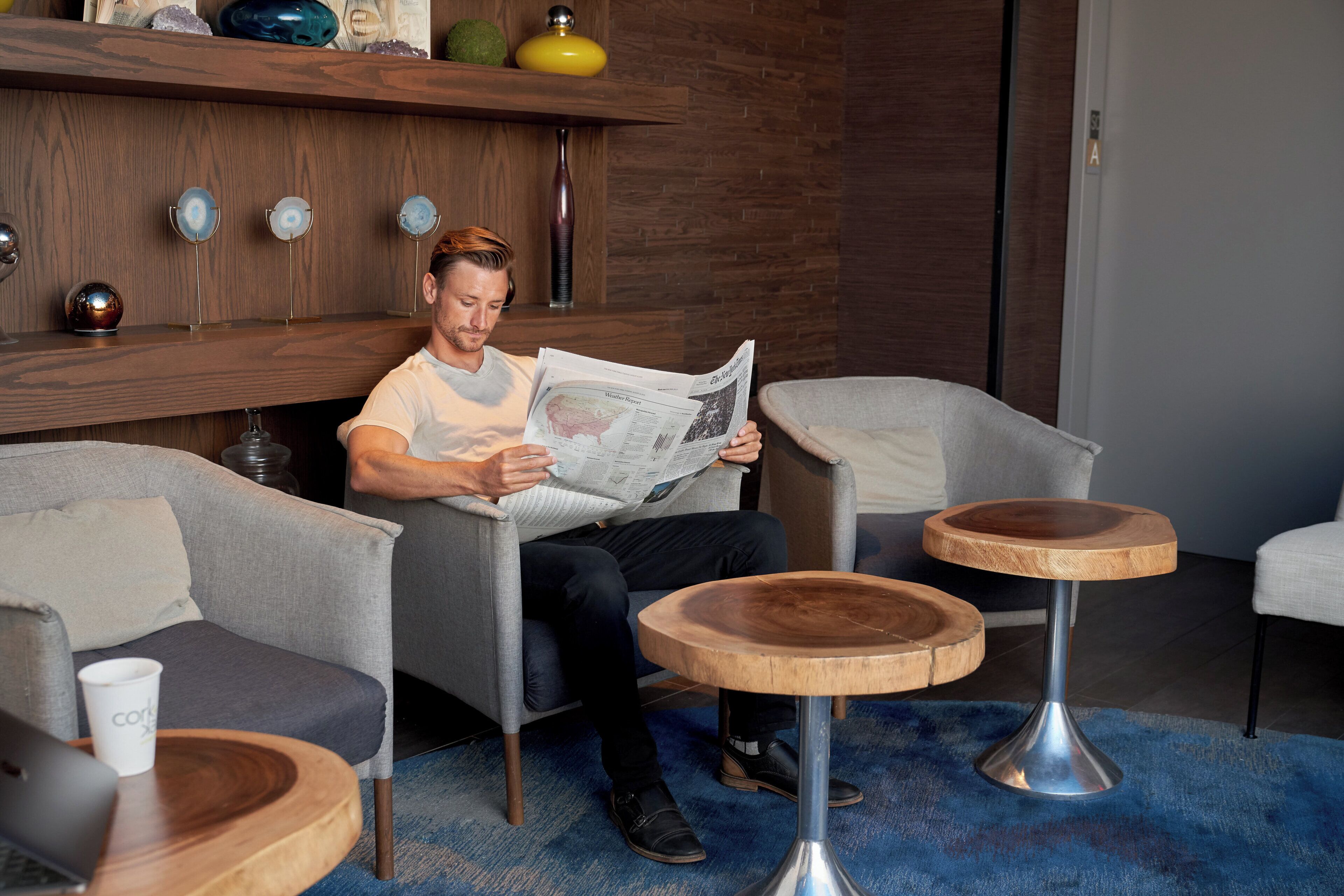 Un hombre leyendo un periódico en un moderno área de descanso con mobiliario de madera y alfombra azul.