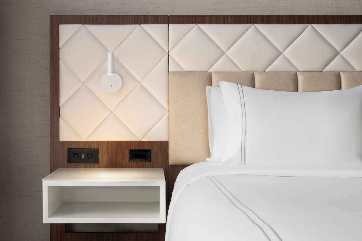 A hotel room with a modern headboard, a bedside table, and a wall-mounted light fixture.