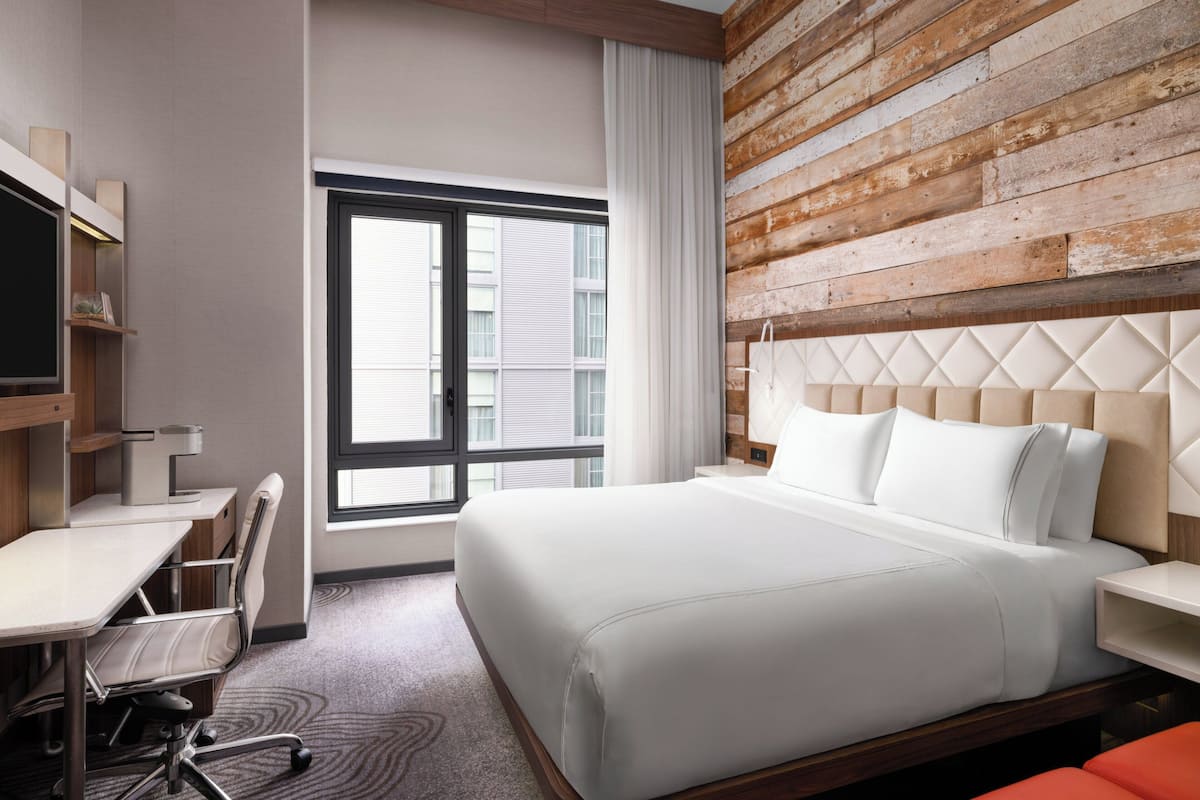 A hotel room with a bed, desk, chair, and a large window.