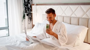 A person in a white robe sitting on a bed with a laptop and a green drink, reading a newspaper. - EVEN Hotel New York - Times Square South by IHG (New York)