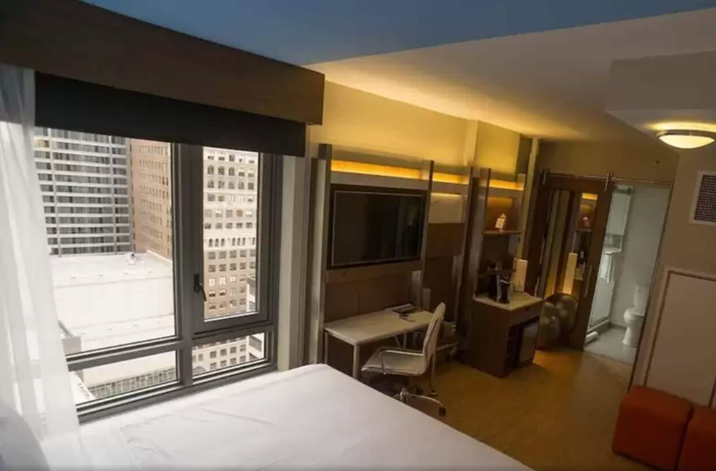 Une chambre d’hôtel comprenant un lit, un bureau, une chaise, une petite table, un miroir et une fenêtre donnant sur des immeubles.